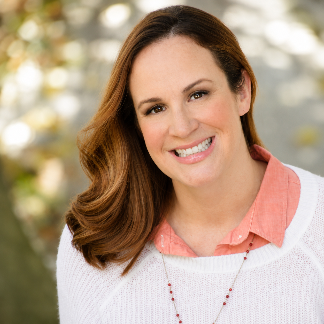 A woman with light skin and brown hair smiling outdoors, wearing a pink collared shirt and white sweater.