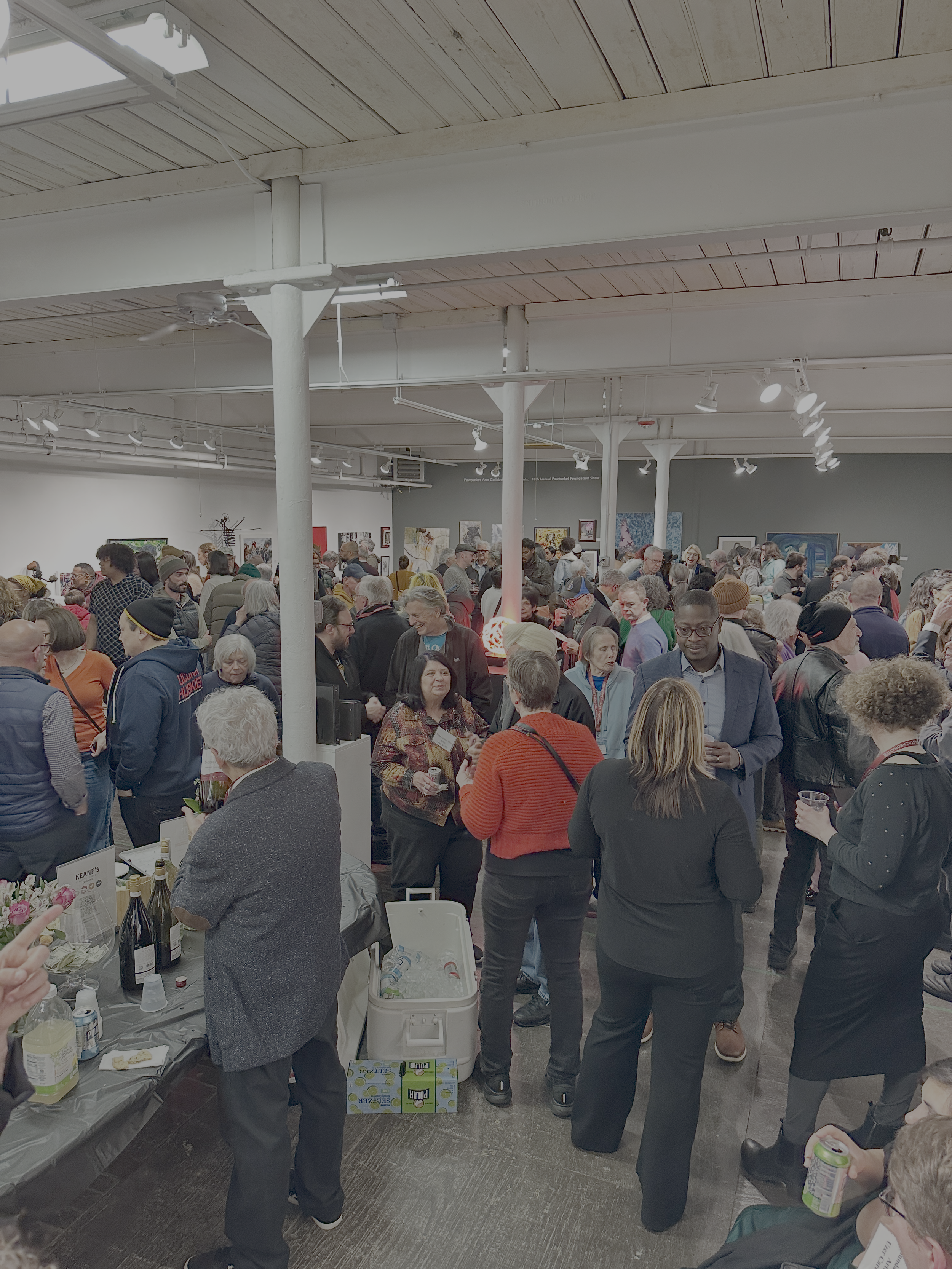 A crowded indoor event with many people browsing booths and tables, some holding cups or bottles, in a gallery or exhibition space with spotlights and artwork on the walls.
