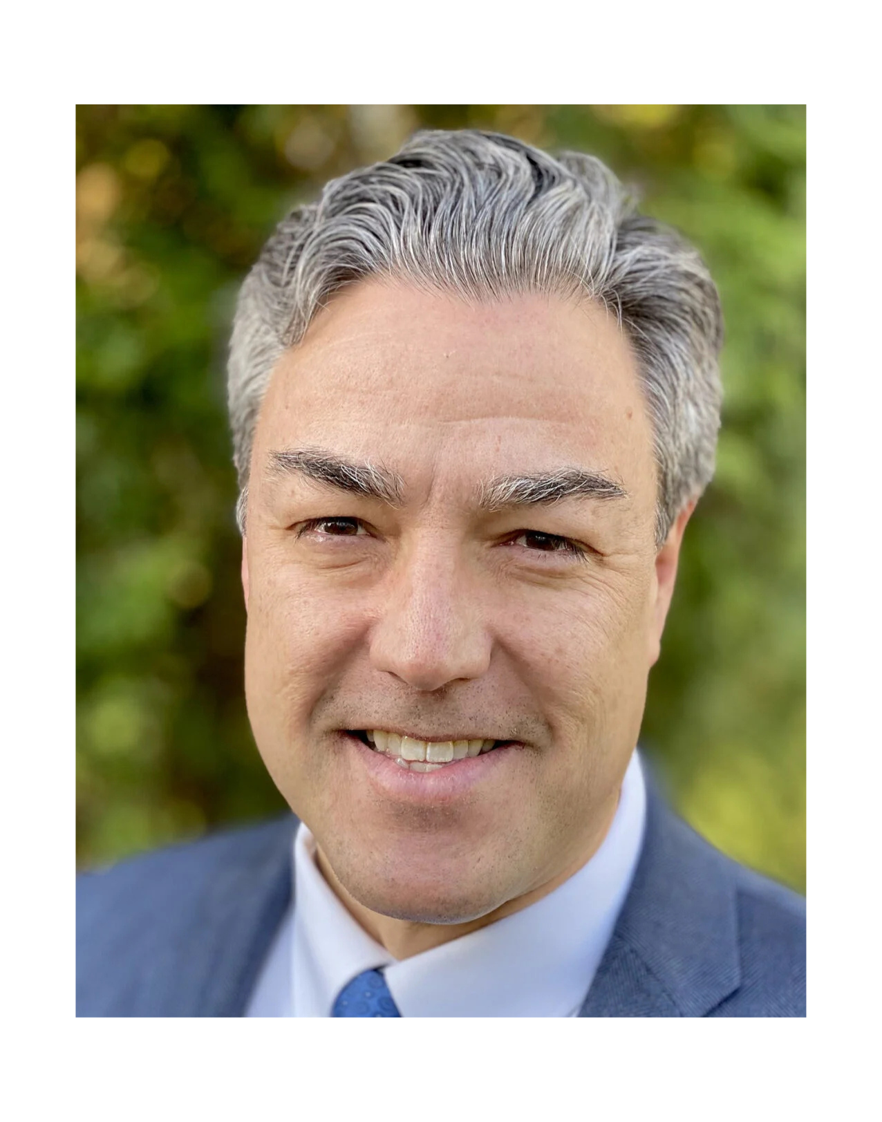 Close-up of a smiling middle-aged man with gray hair, wearing a blue suit, white shirt, and blue tie, outdoors with blurred green background.