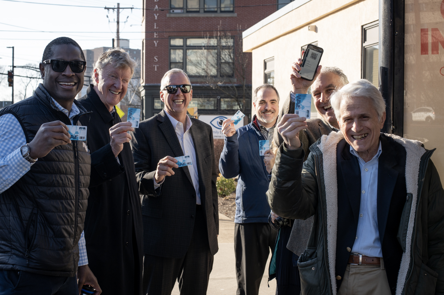 A group of six smiling men standing outdoors, holding up augmented reality or digital tickets. Some are wearing sunglasses, and they are standing in front of a building with a sign and some visible power lines.