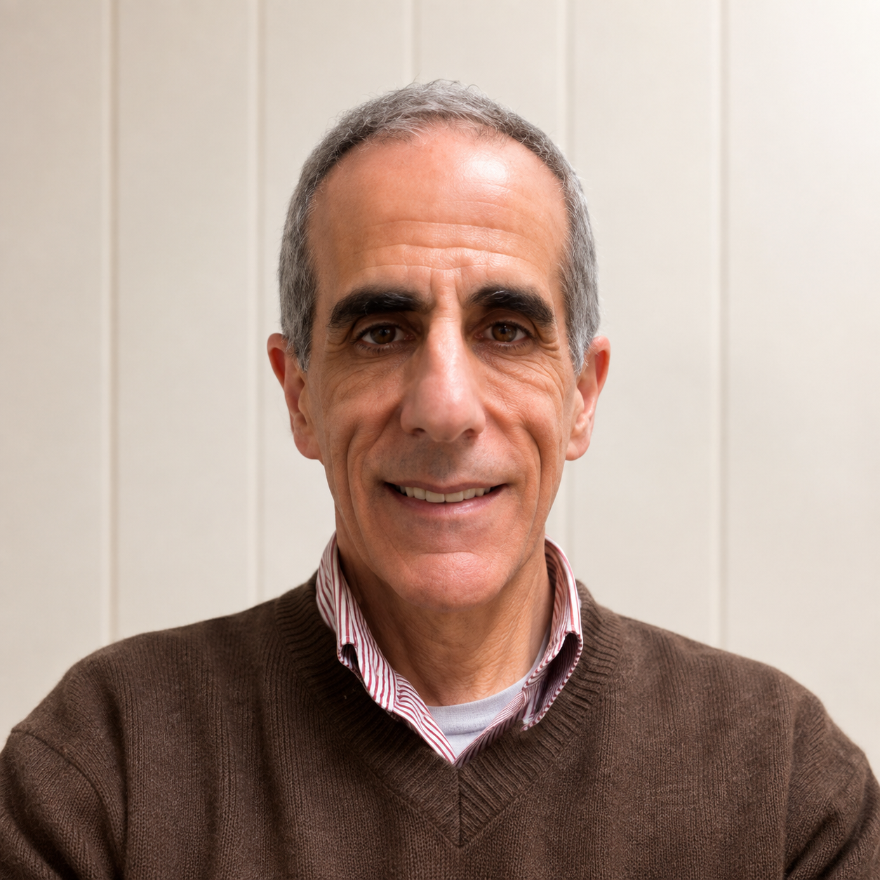 Close-up portrait of a middle-aged man with short gray hair, wearing a brown sweater over a striped shirt, smiling, with a plain light-colored wall in the background.