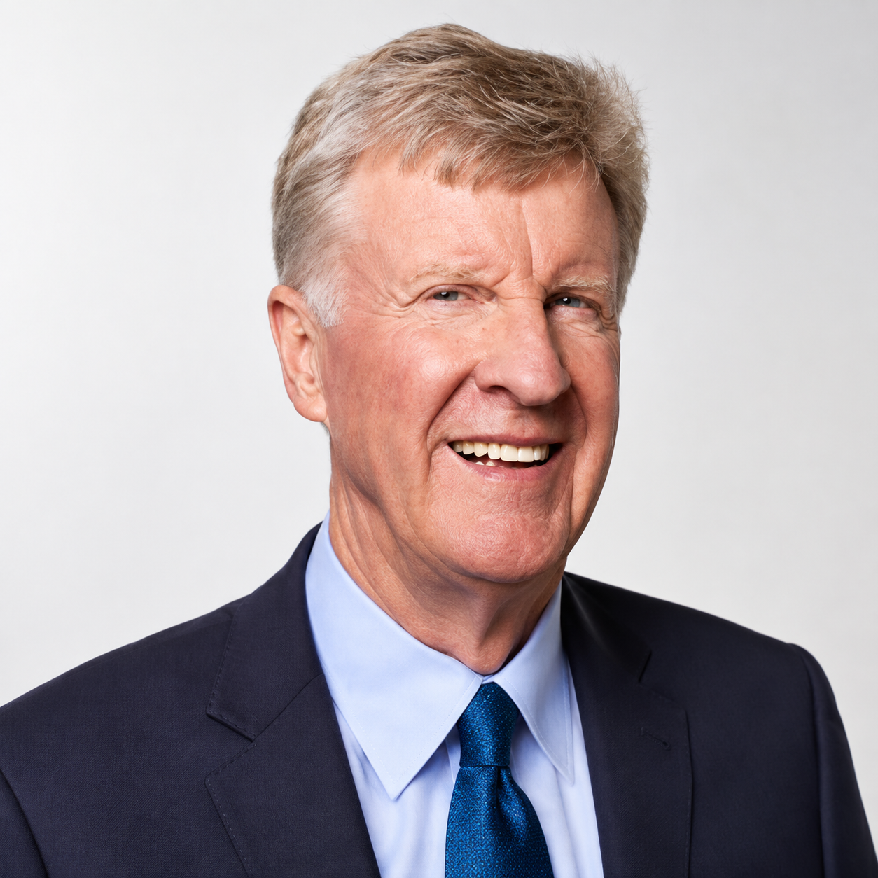Senior man with gray hair smiling, wearing a dark suit, light blue shirt, and blue tie, against a plain white background.