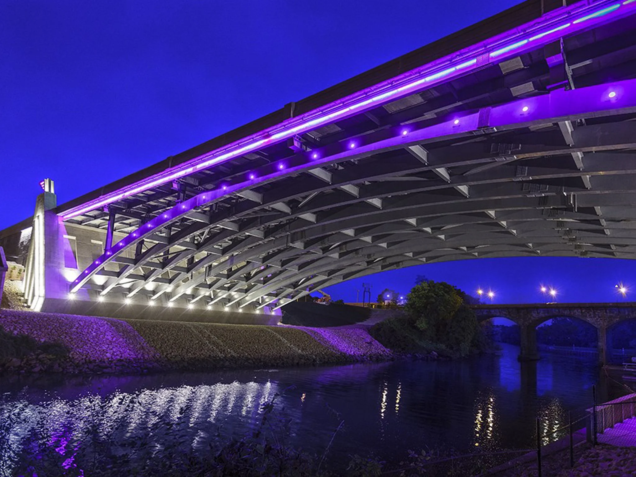 Night view of a bridge illuminated with purple and blue LED lights, reflecting in the water below.