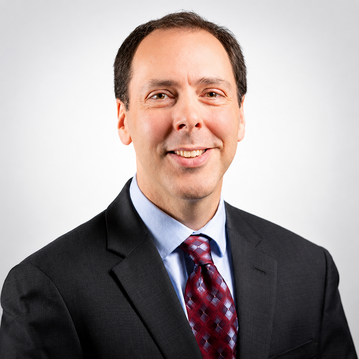 Professional headshot of a smiling man in a black suit, light blue shirt, and patterned red tie, with a neutral gray background.