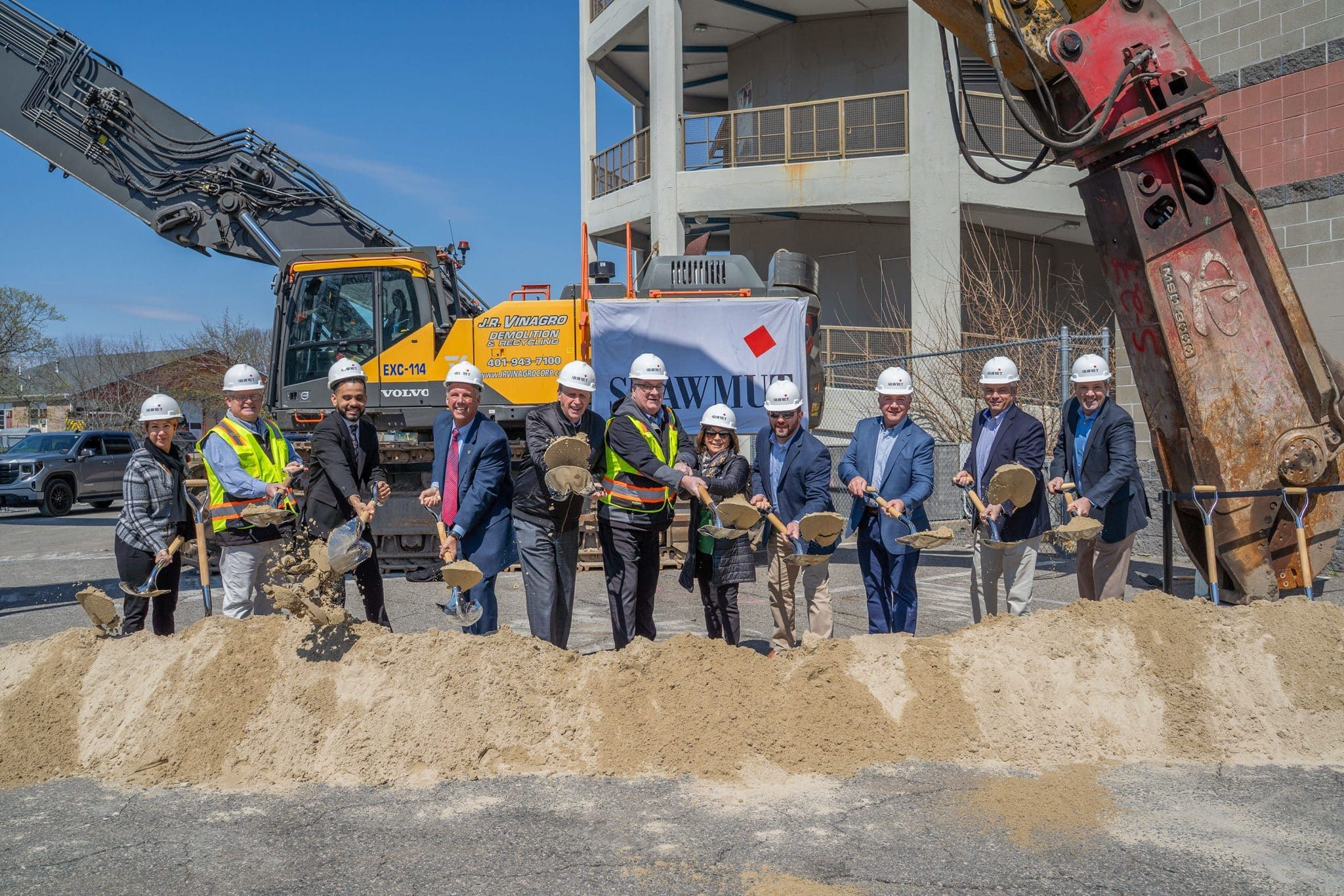 Group of people in construction gear holding shovels at a groundbreaking ceremony in front of construction equipment and a building under construction.