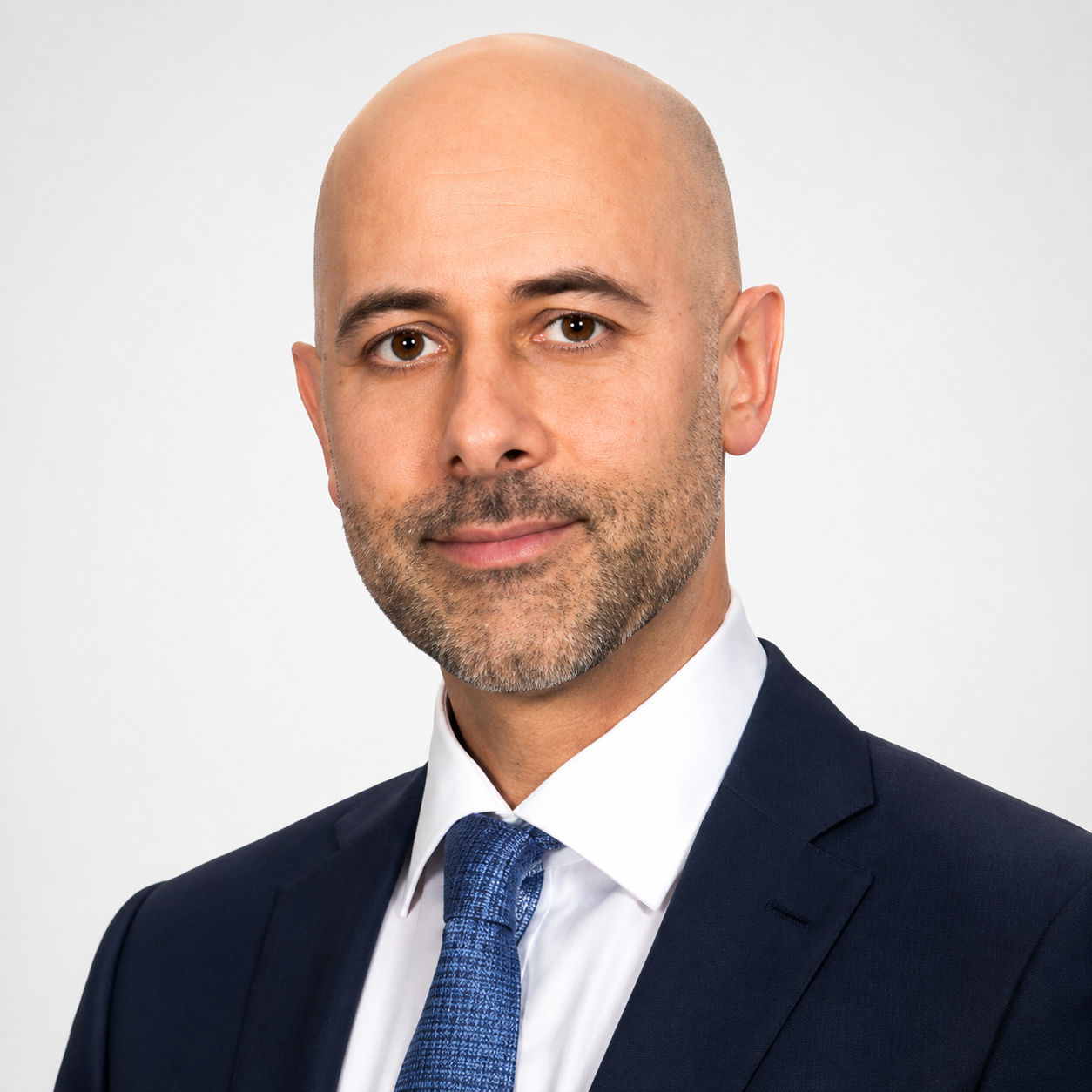 A bald man with a beard, wearing a navy suit, white shirt, and blue tie, posing against a plain white background.
