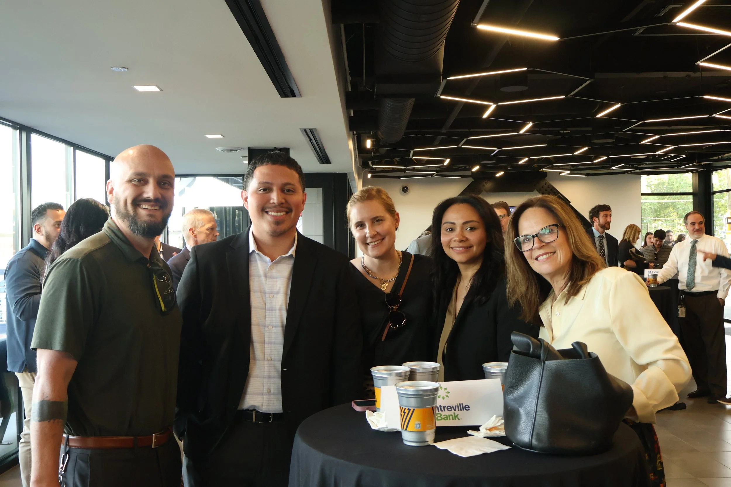 Group of five diverse people smiling at a social event in a modern indoor space with dark ceiling lights and large windows, with other people in the background.
