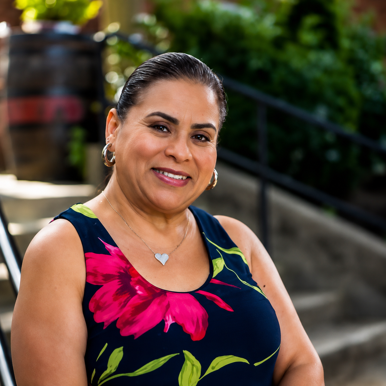 A woman smiling outdoors wearing a sleeveless navy dress with pink and green floral pattern, silver hoop earrings, and a silver heart necklace.