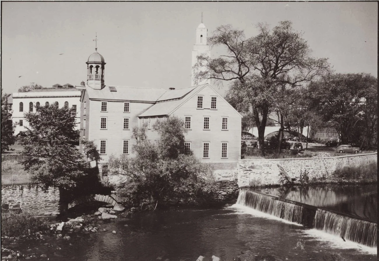 A black and white photo of historic buildings with a river in the foreground, trees, and parked cars in the background.