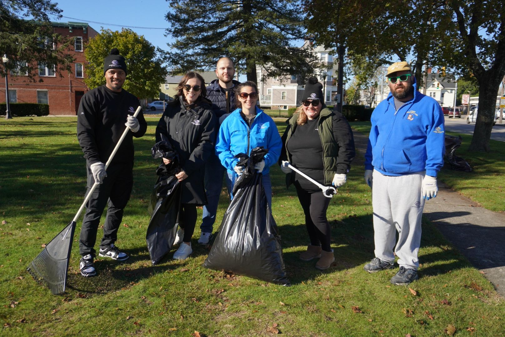 Group of six people participating in a park cleanup, holding trash bags and litter pickup tools, standing on grass with trees and buildings in the background on a sunny day.
