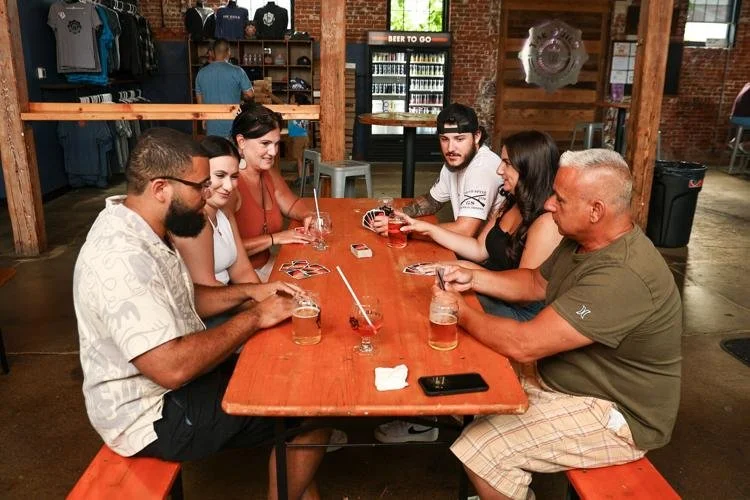 Group of six people playing cards at a wooden table in a brewery or pub with drinks.