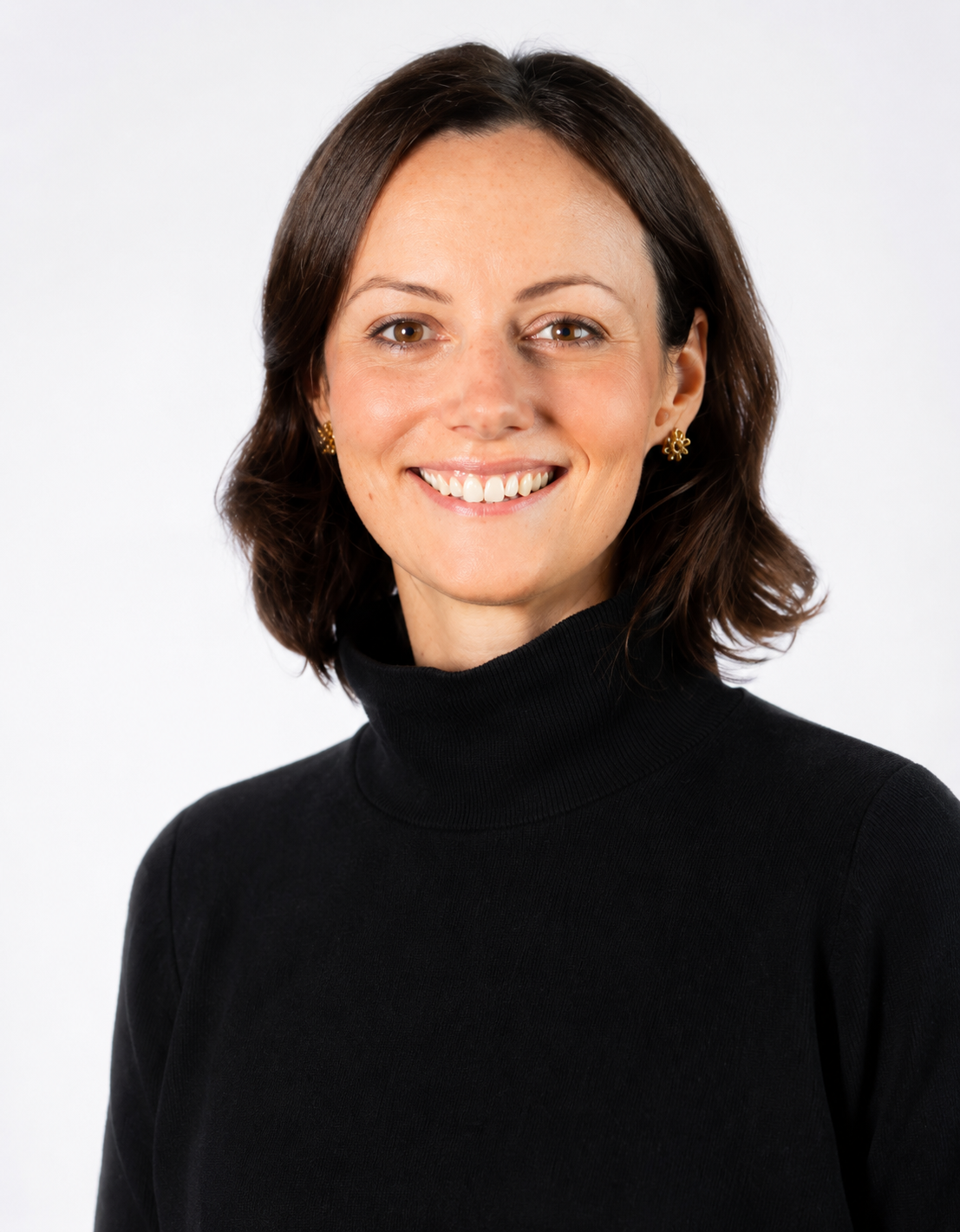 Portrait of a woman with dark brown hair, wearing a black turtleneck and gold earrings, smiling at the camera against a plain white background.