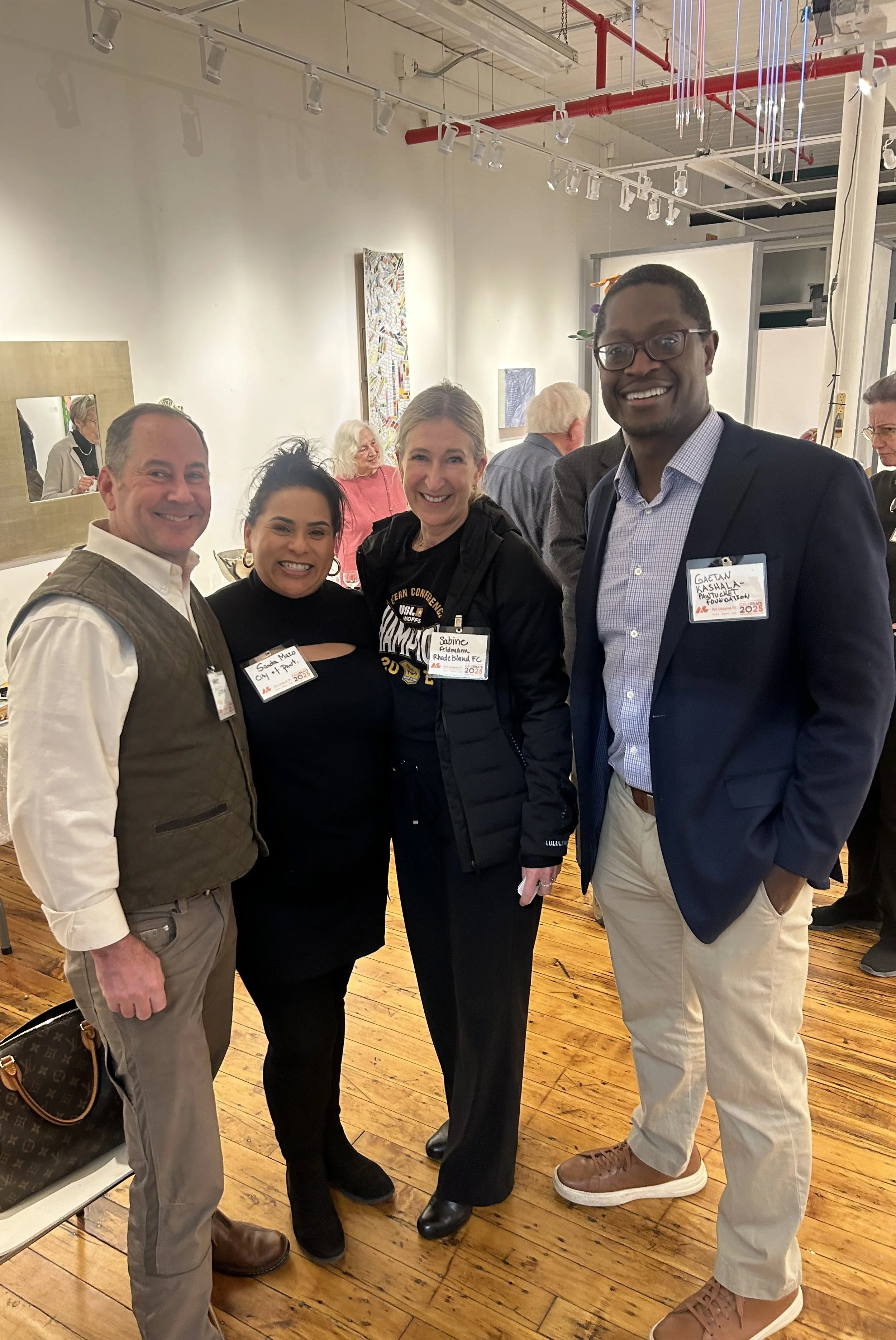 Four people standing indoors, smiling at the camera. They appear to be at an event or gathering with artwork on the walls and other people in the background. There are two women and two men, all wearing semi-formal clothing and name tags.