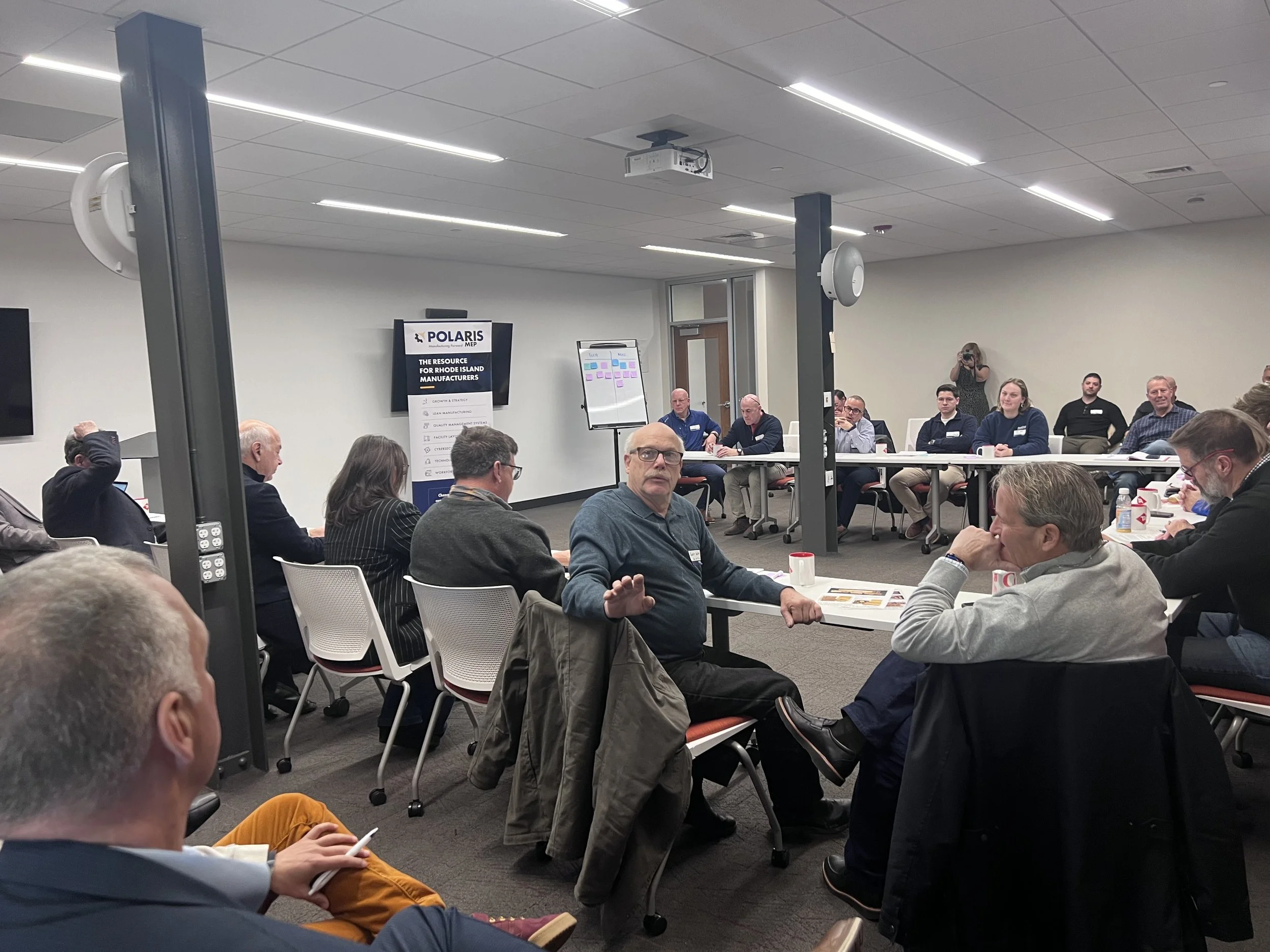 A group of people attending a conference or meeting in a modern conference room. Some are seated at tables, listening, and some are taking notes. There is a presentation screen on the wall displaying a sign for Polaris, a manufacturing resource. A woman is taking photos in the background.