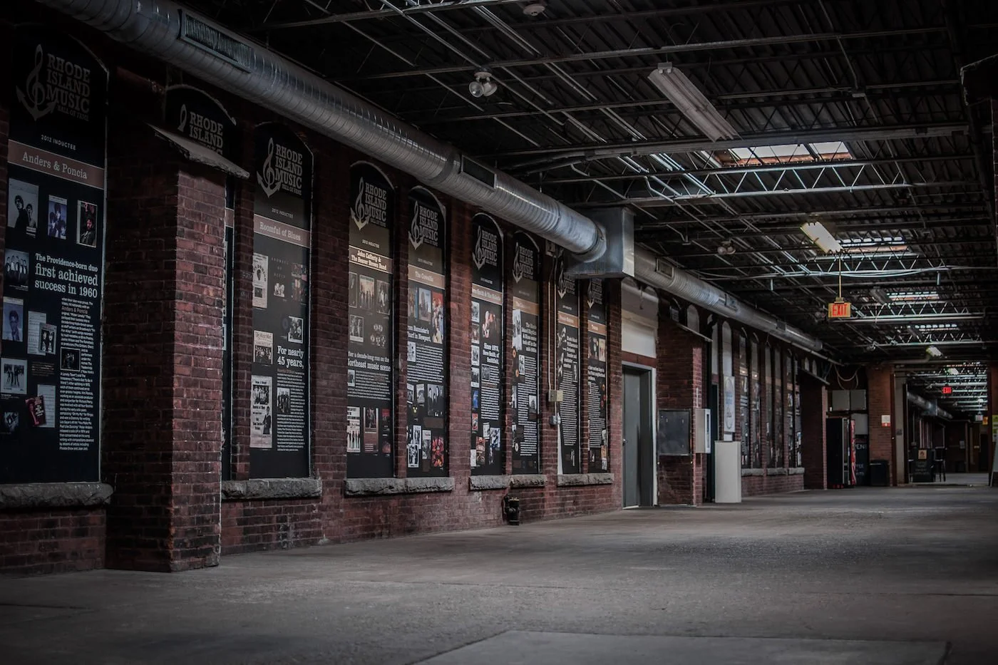 Empty hallway with brick walls and informational posters about Rhode Island music history, industrial ceiling with ductwork, and vending machines at end.