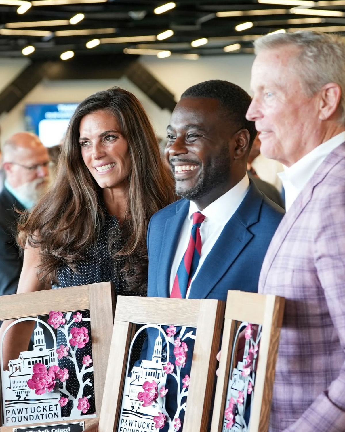 Three people standing behind a table with awards, smiling at an event. Women and men dressed in formal attire, with awards featuring the Pawtucket Foundation logo and pink blossoms in front of them.