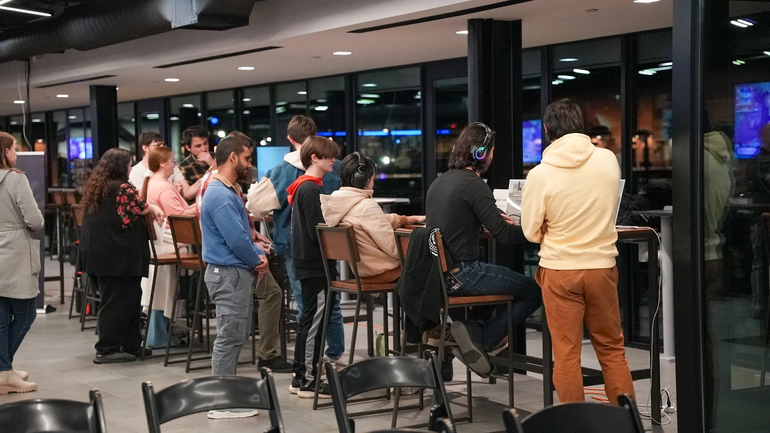 A group of diverse people, including men and women, standing and sitting in a line at an indoor gaming or internet cafe at night, with some wearing headphones and playing games on computers through large glass windows.