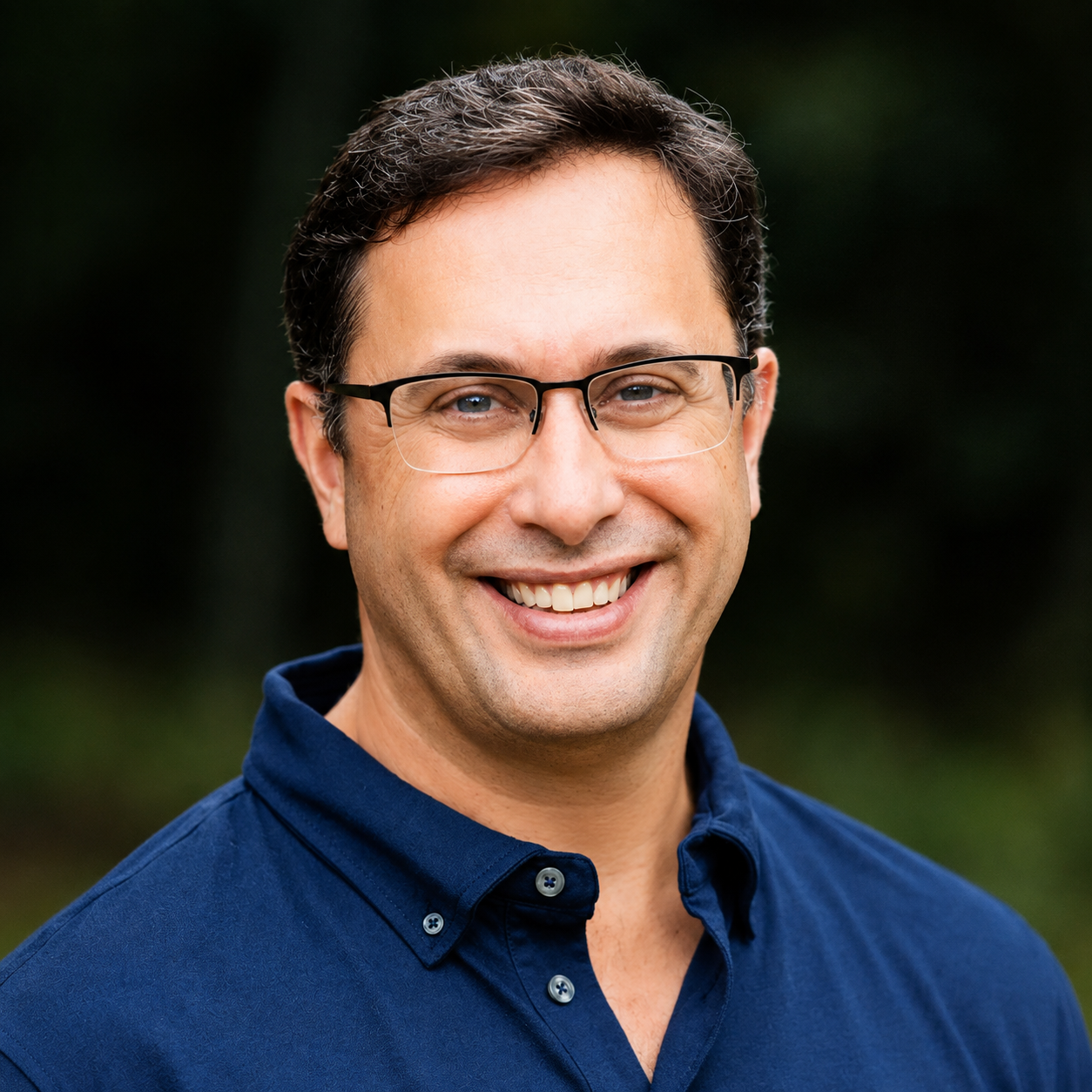 Headshot of a smiling man with dark hair, glasses, and wearing a navy blue button-up shirt, photographed outdoors with blurred greenery background.