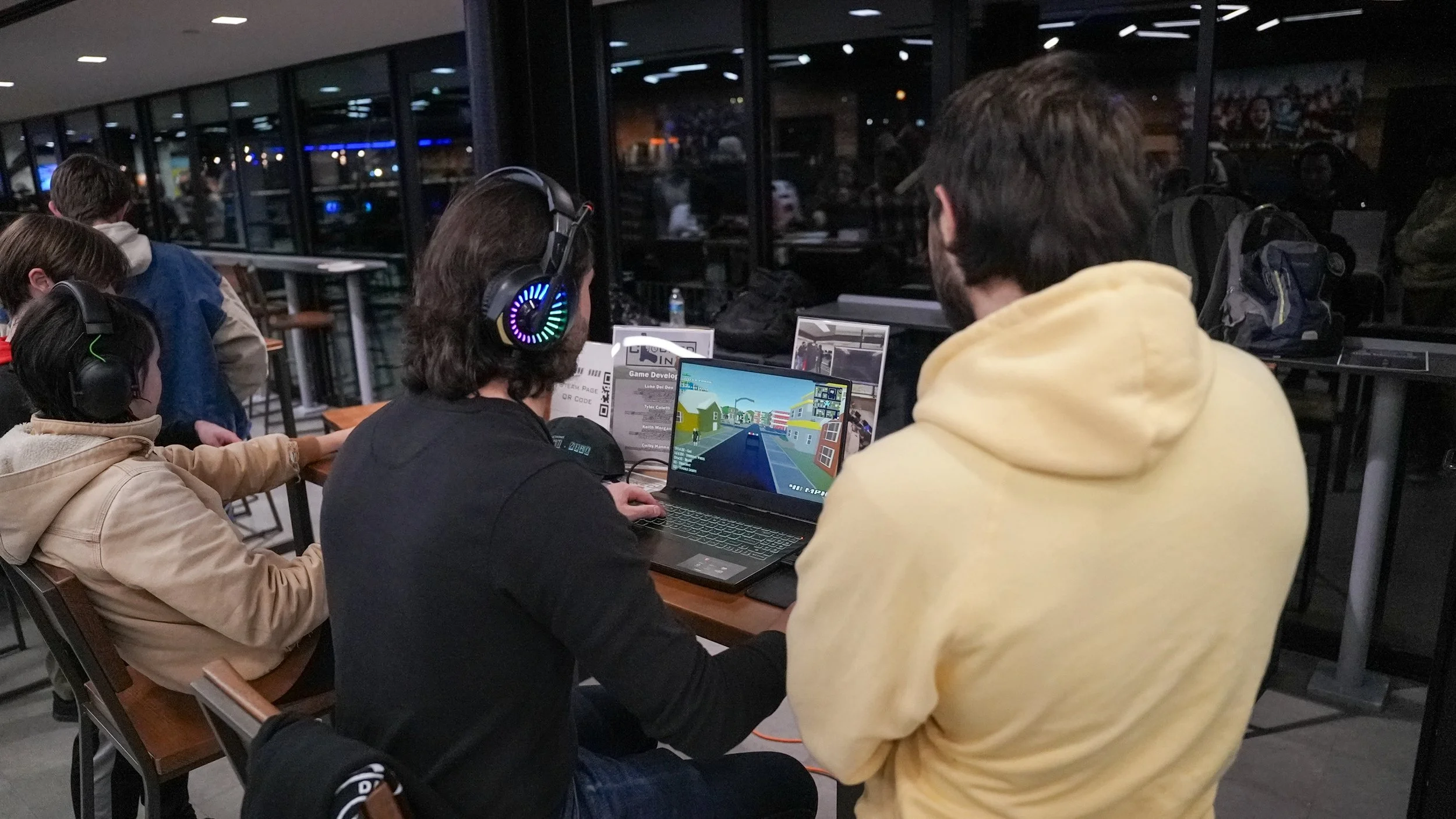 People gathered in a dimly lit indoor space, some wearing headphones, playing video games on laptops and computers, with a view of a city street outside through large windows.