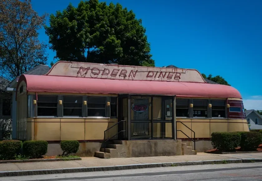 An old, rusty building with a sign reading 'Modern Diner' on the roof, front steps leading up to a glass door, surrounded by a sidewalk and small bushes, with trees and a blue sky in the background.