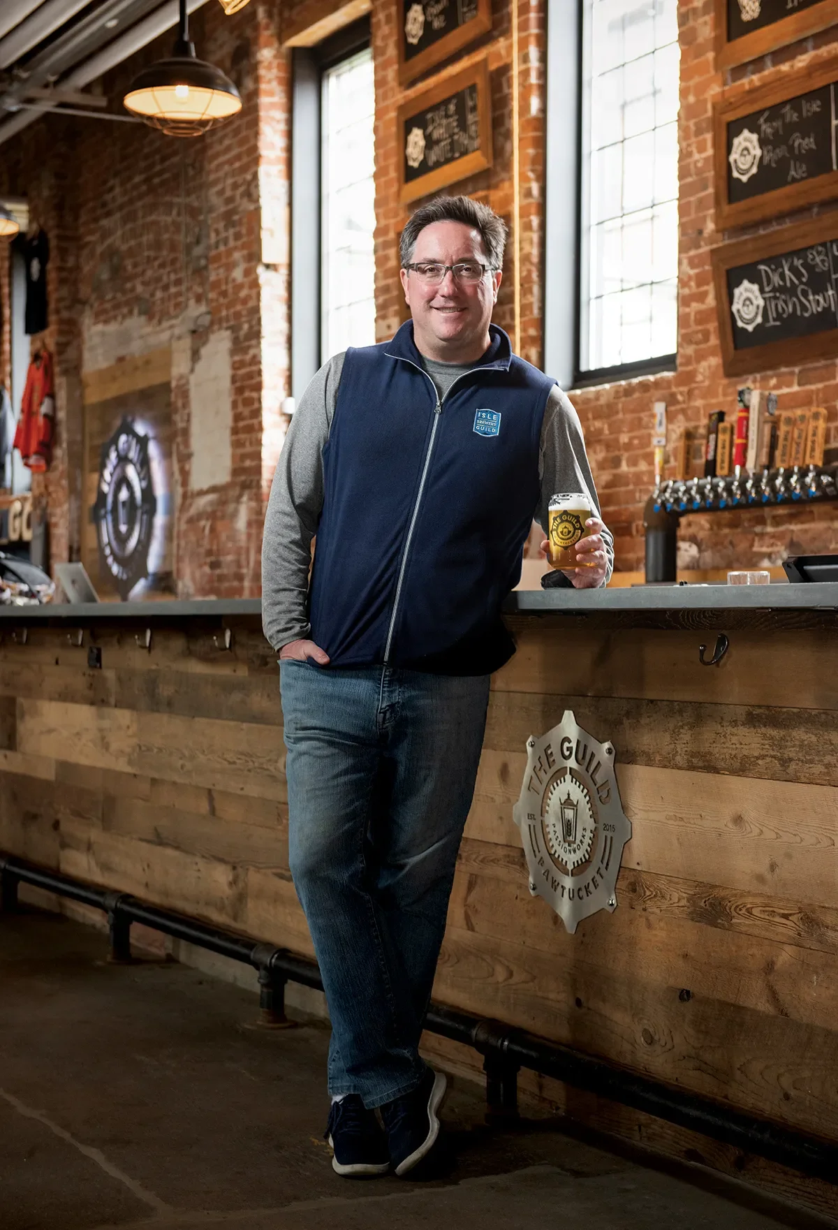 A man with glasses, wearing a blue vest and jeans, stands at a bar holding a glass of beer, inside a rustic brewery with exposed brick walls and large windows.