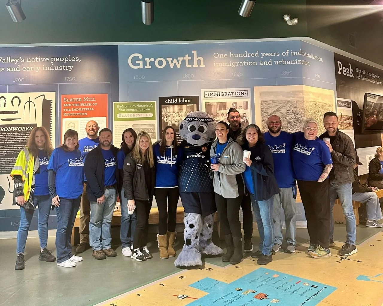 Group of people standing together indoors, posing with a costumed mascot in front of a large informational wall exhibit at a museum or historical site.