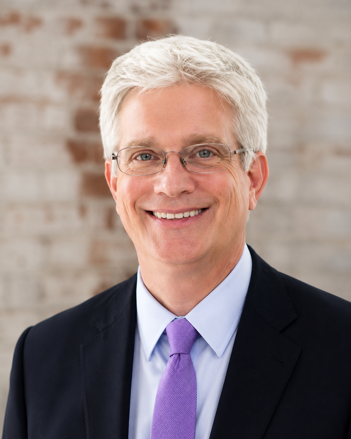 A middle-aged man with gray hair and glasses, wearing a dark suit, light blue shirt, and a purple tie, smiling against a blurred brick wall background.