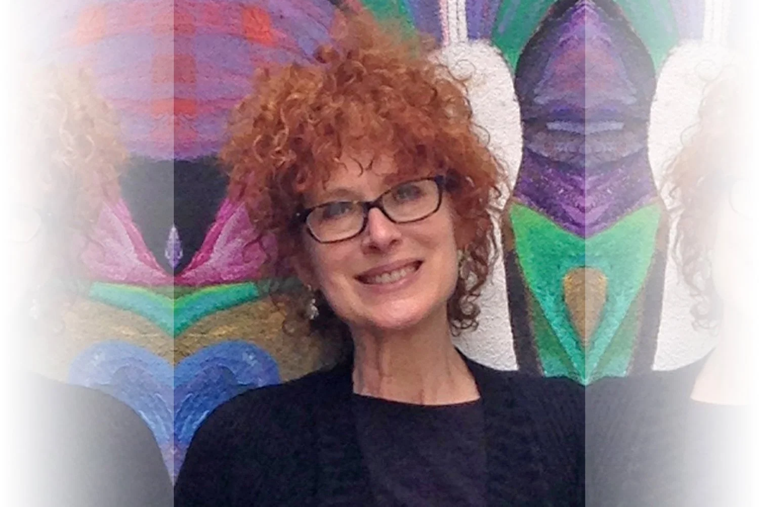 A woman with curly red hair and glasses smiling in front of a colorful abstract painting.