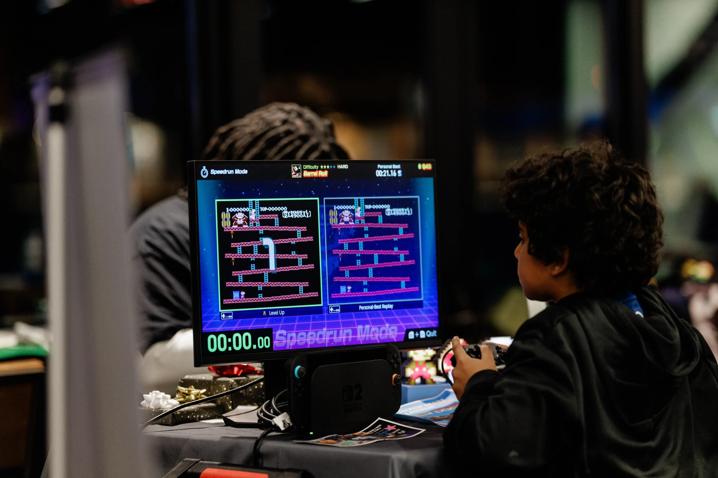 Child playing an arcade game called 'Donkey Kong' at an event or arcade, with a monitor showing gameplay in the foreground.