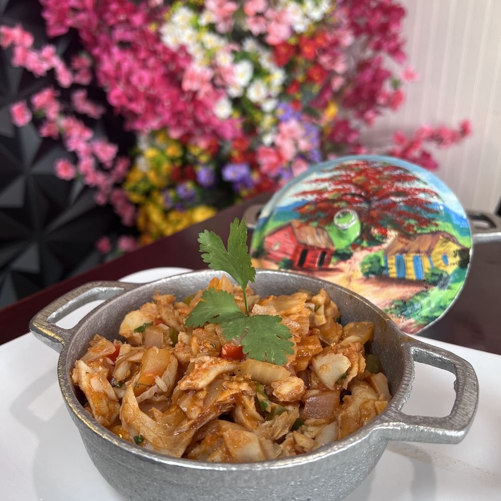 A small silver dish of cooked shredded chicken with vegetables, garnished with cilantro, in front of a colorful painting and a large bouquet of pink, purple, white, red, and yellow flowers in the background.