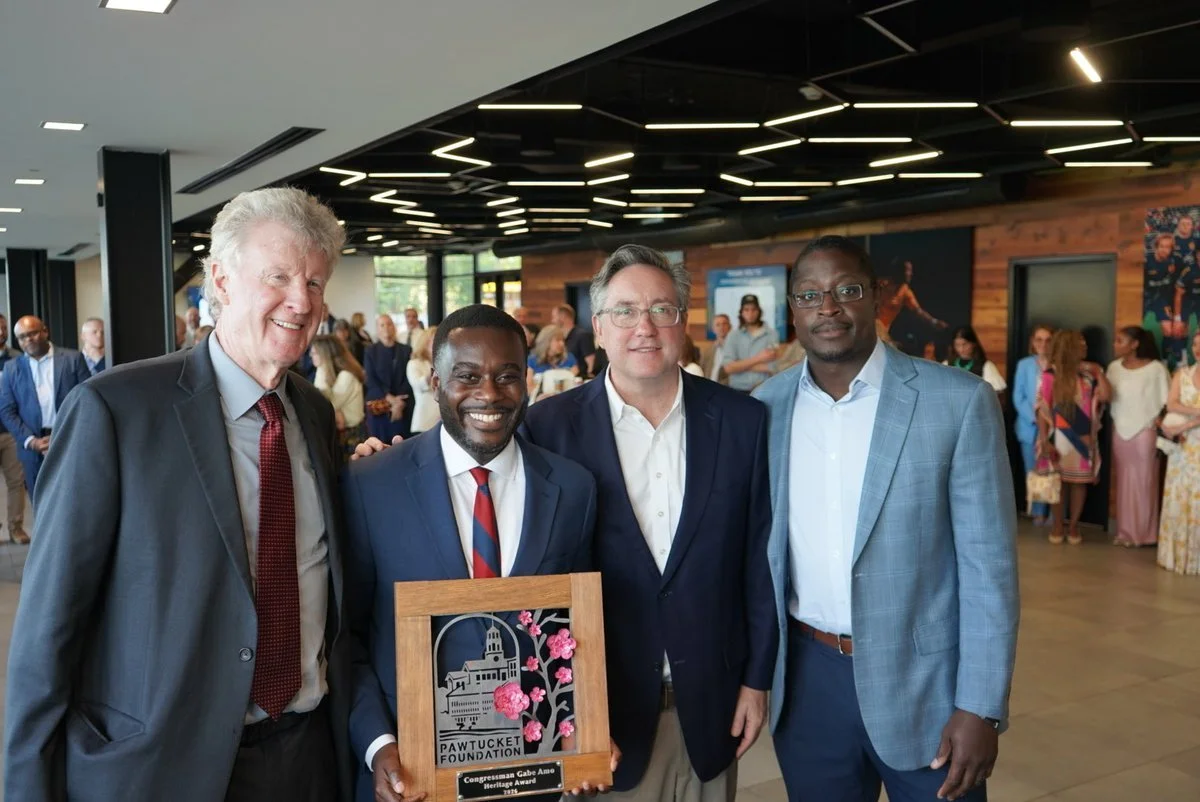 Four men in suits standing in a modern event space, with a group of people in the background. One man holds a plaque with a wooden frame and a design of a building and pink flowers, reading 'Pawtucket Foundation'.