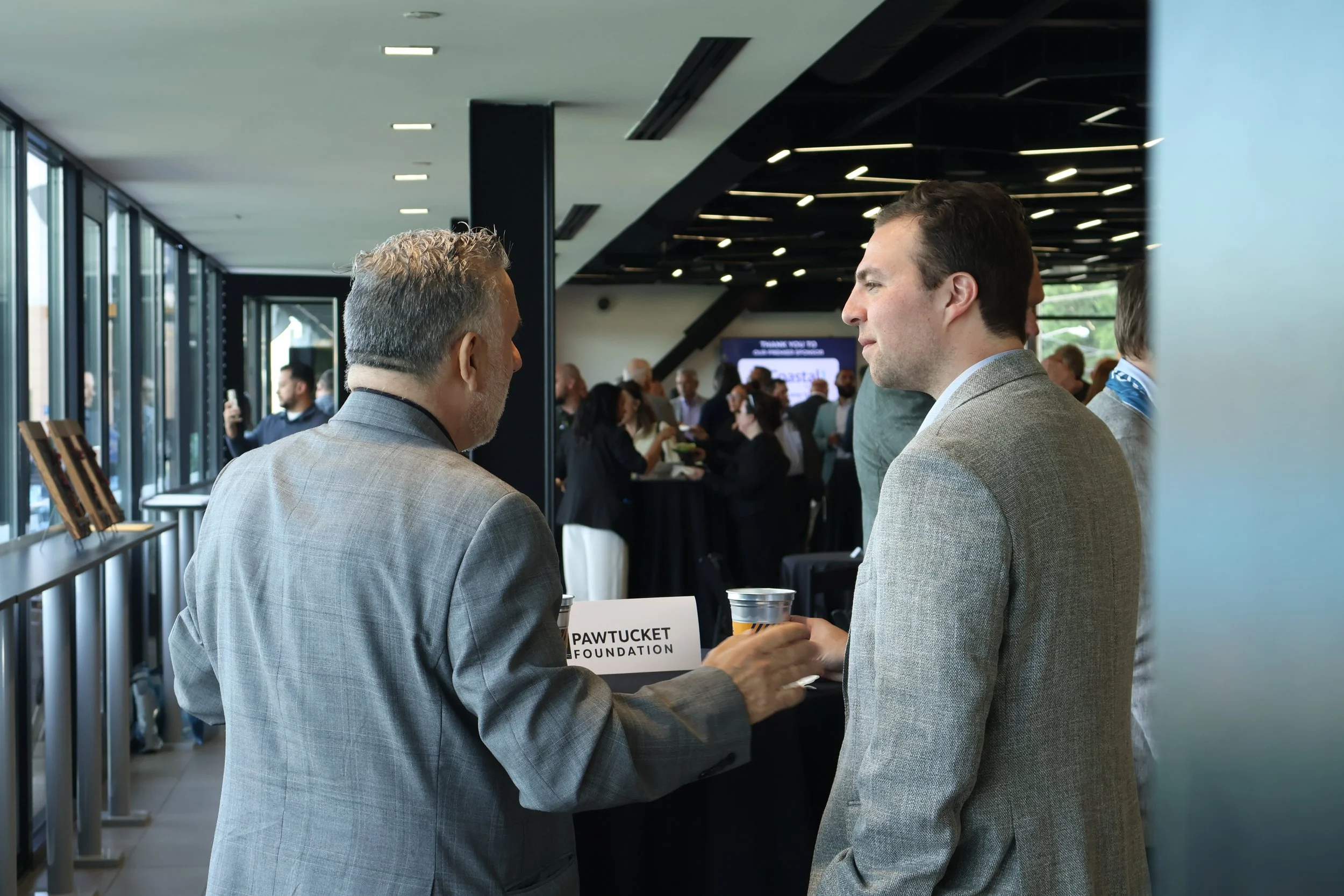 Two men in business suits engaged in conversation at a professional event, holding drinks, with a crowded modern conference space in the background.