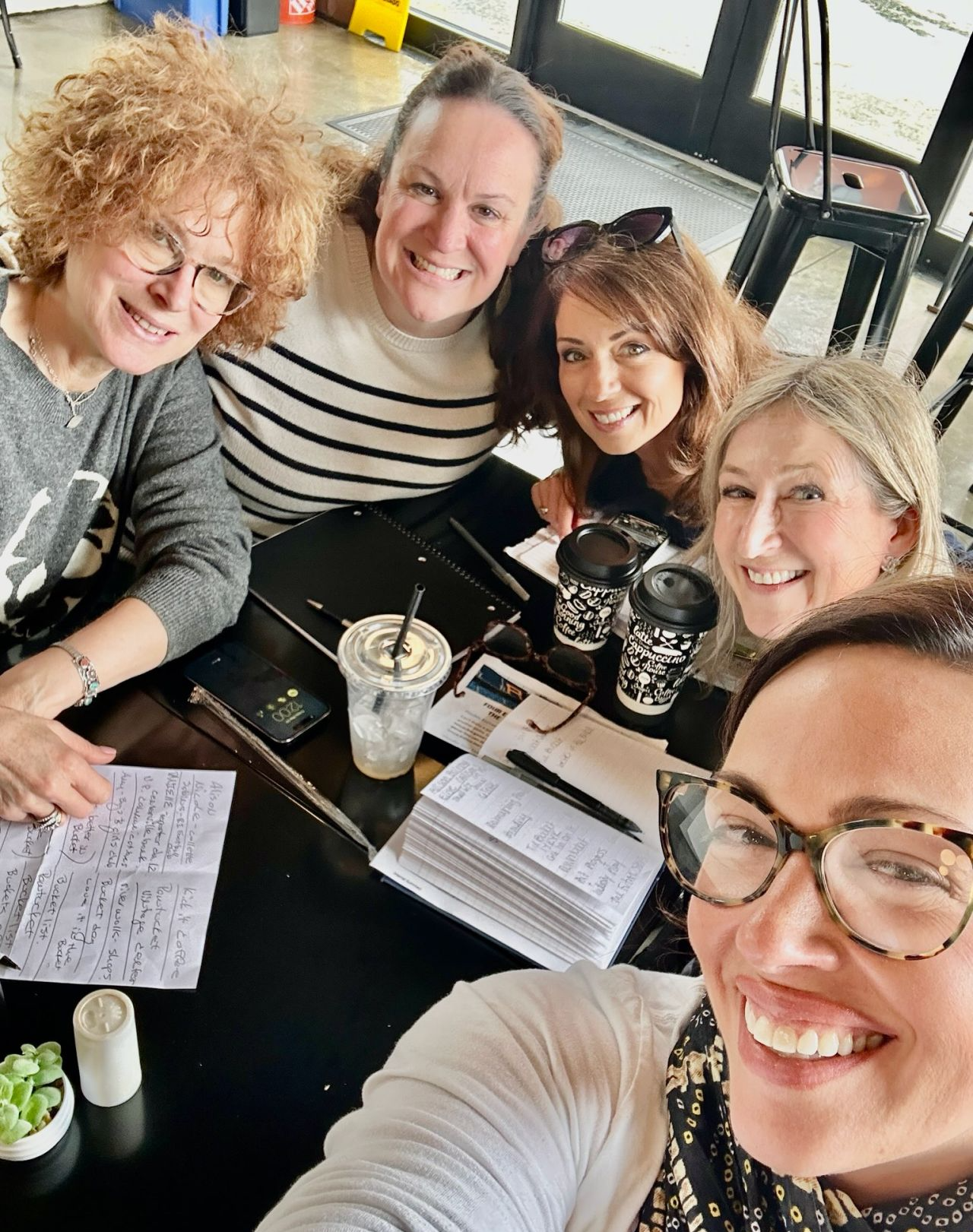 A group of six women smiling and taking a selfie at a table with notebooks, coffee cups, and a drink in a cafe with glass doors.