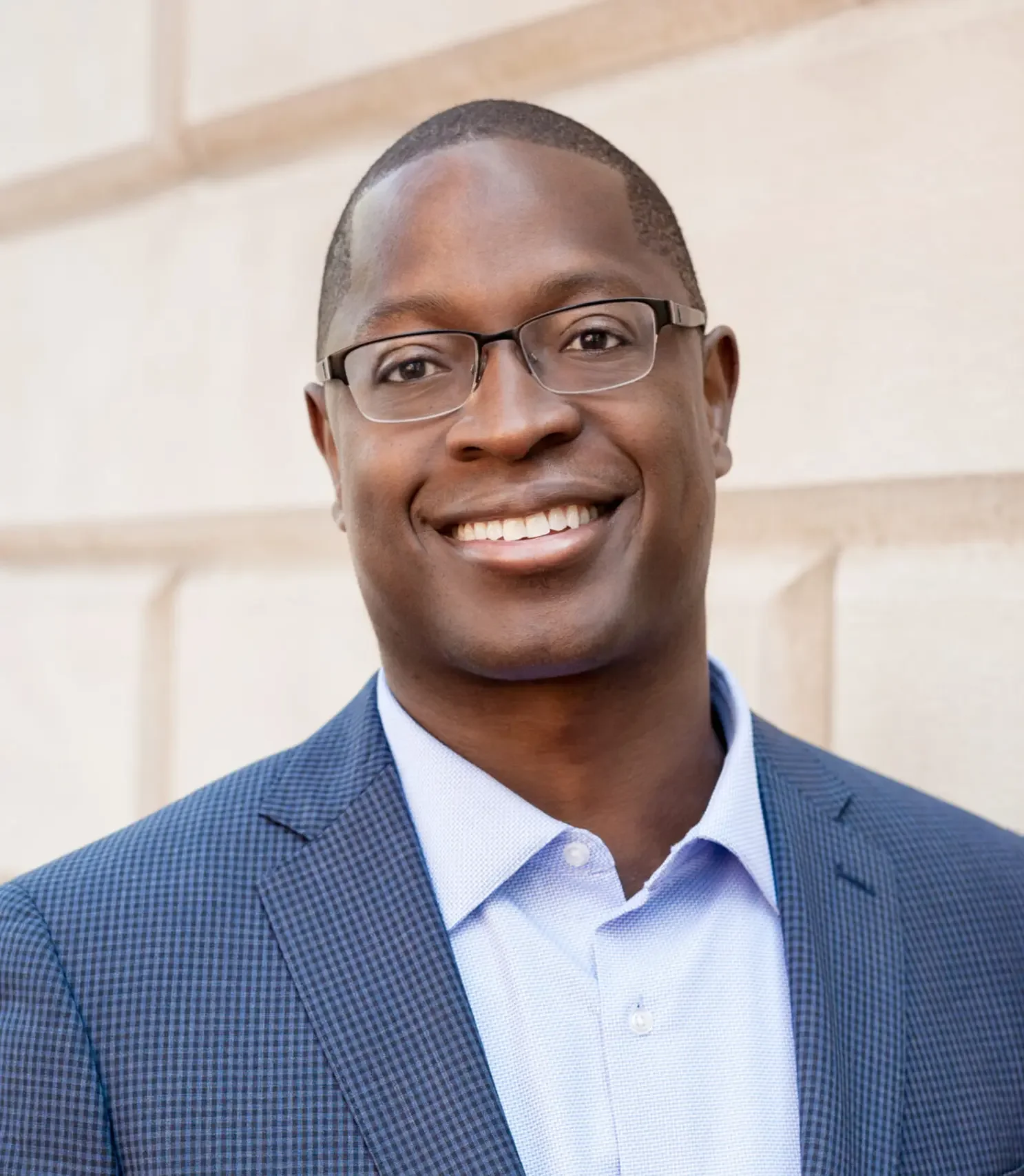 Headshot of a smiling man wearing glasses, a blue checkered suit jacket, and a light blue button-up shirt.