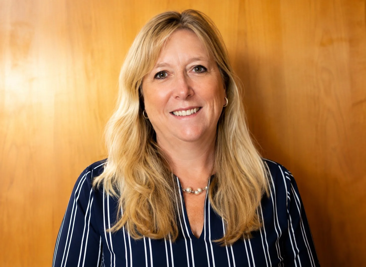 A woman with blonde hair smiling, wearing a navy blue and white striped blouse, a pearl necklace, and hoop earrings, standing against a wooden background.