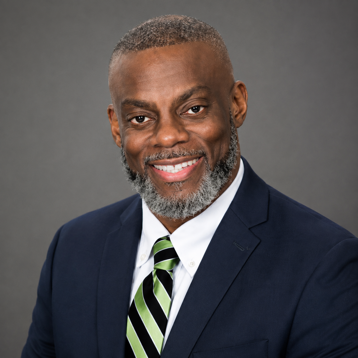 Portrait of a smiling African American man with short gray hair and a beard, wearing a navy suit, white shirt, and green-striped tie against a gray background.
