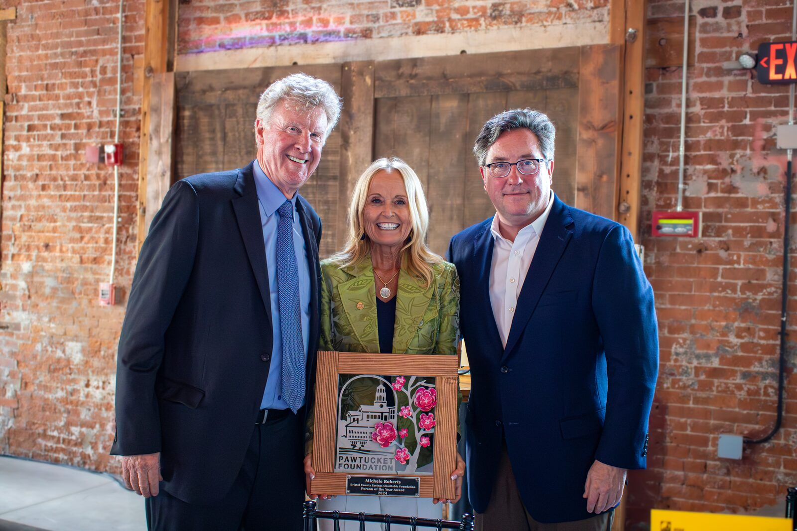 Three people, two men and one woman, smiling and standing indoors against a brick wall; the woman holds a framed award with a cityscape and pink flowers, and a label reading 'Pawtucket Foundation' and 'Michele Roberts'.