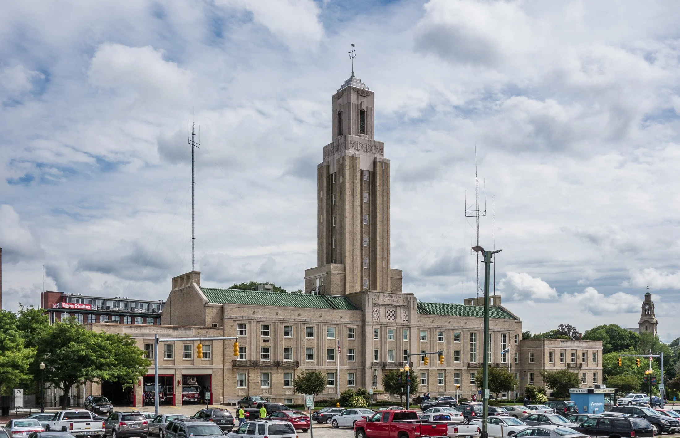 A large, ornate city hall building with a tall clock tower topped by a weather vane. The building has a green roof and intricate stonework, with several surrounding trees and a busy parking lot in front, under a partly cloudy sky.