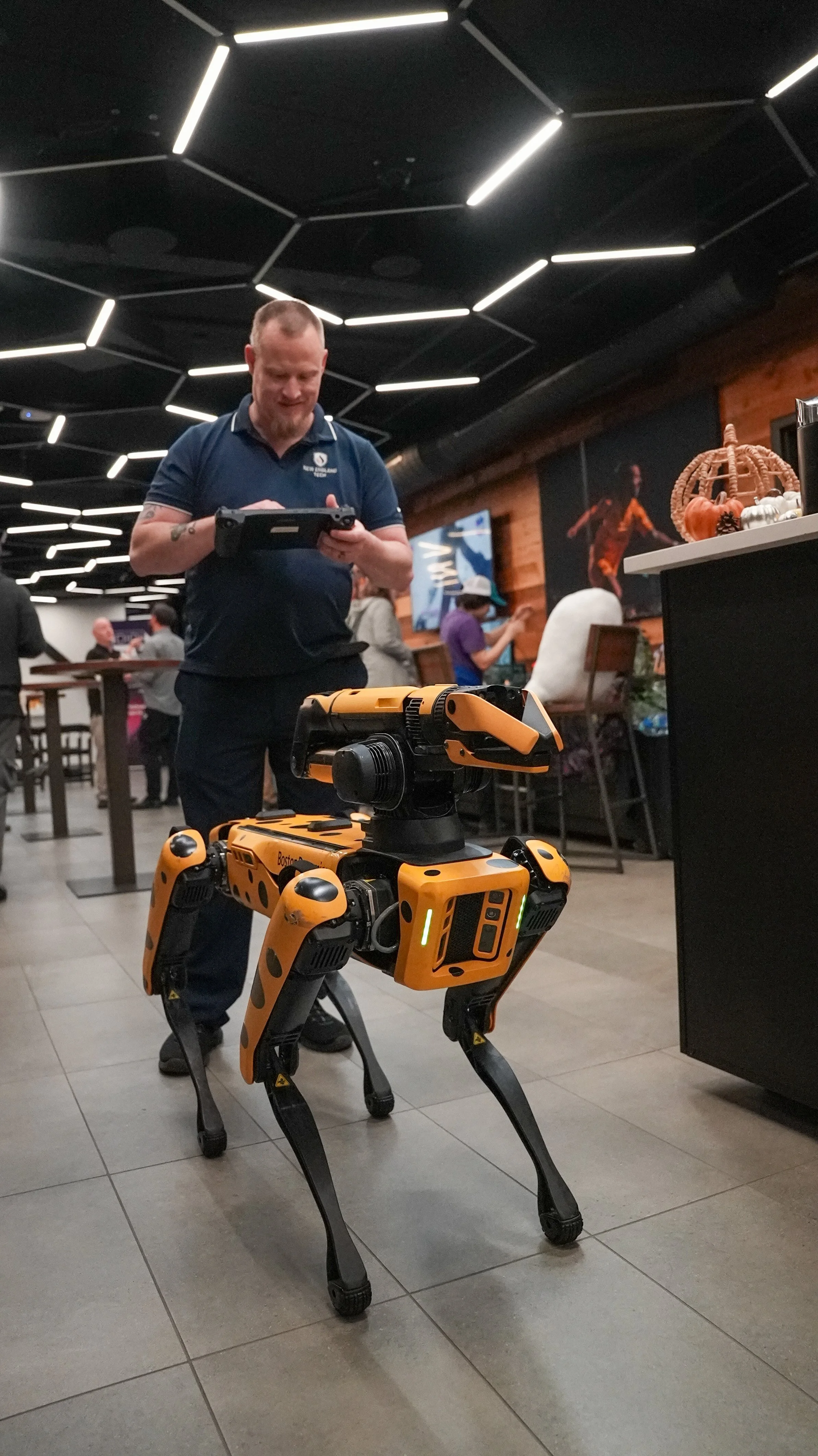 A humanoid robot with a four-legged design, orange and black body, standing on a tiled floor in a restaurant or cafe. A man is looking at a tablet in the background, and there are people and decorations in the surroundings.