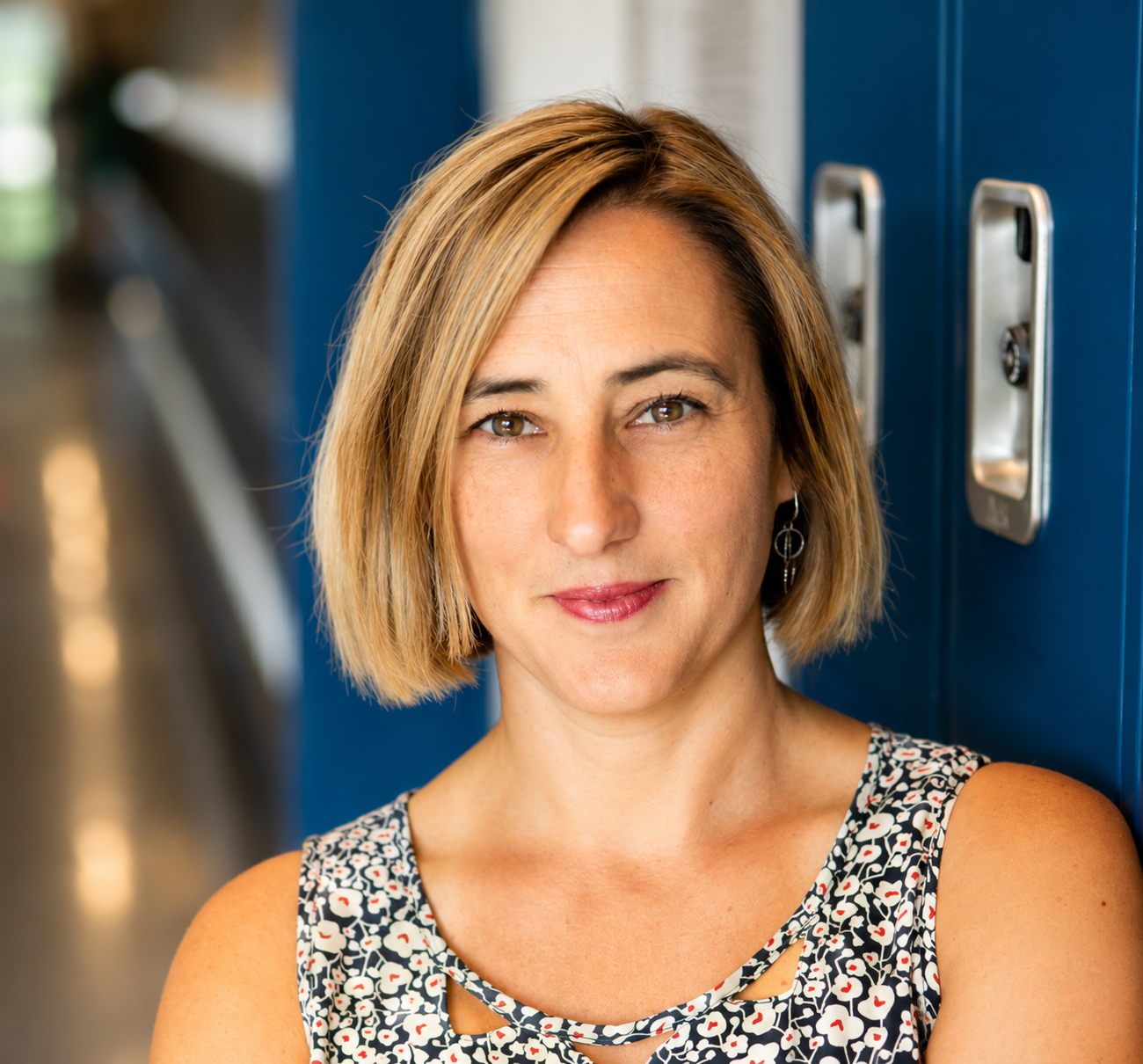 A woman with light brown hair and brown eyes standing in front of blue lockers in a school hallway.