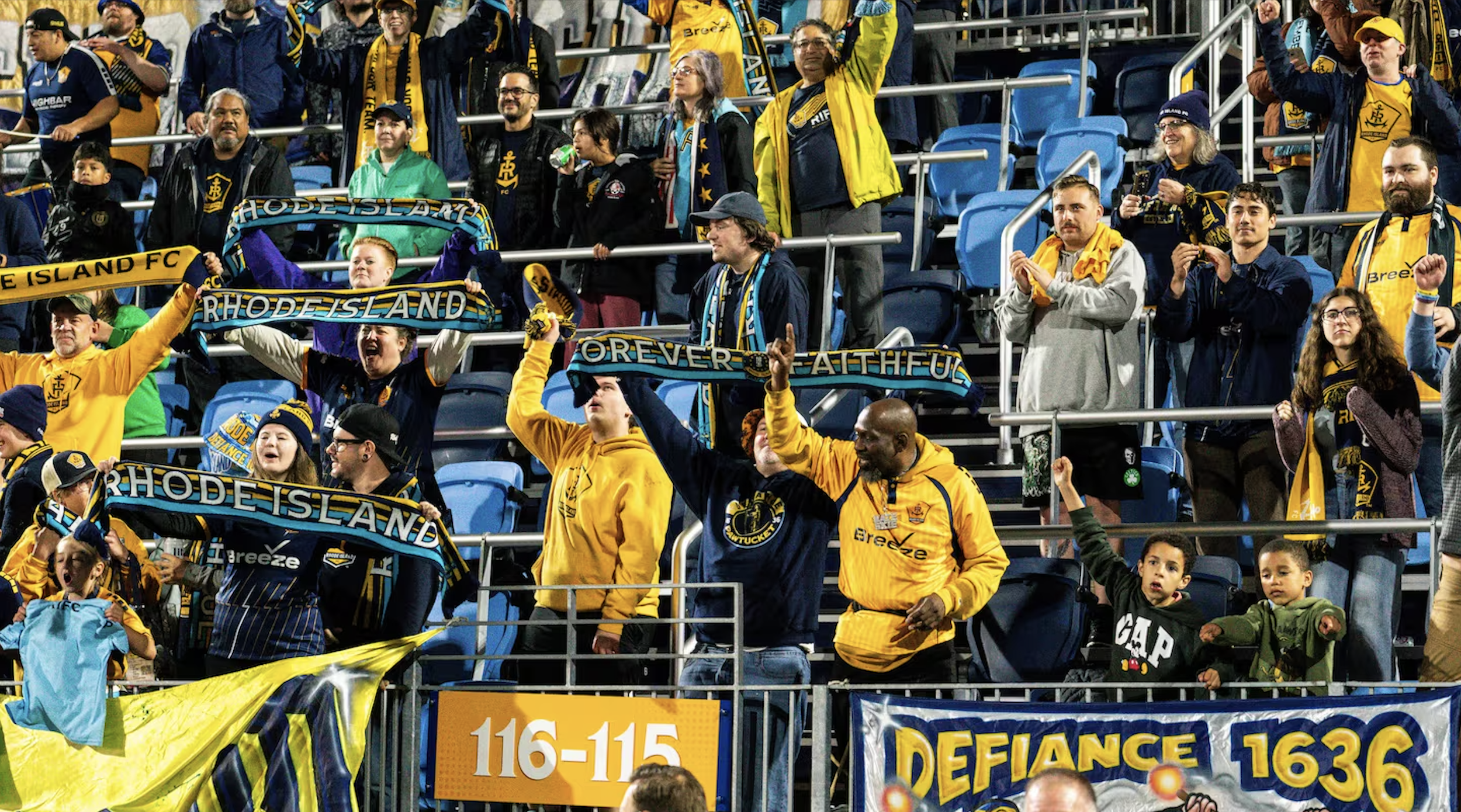 A group of soccer fans at a stadium, wearing yellow and navy jerseys and scarves supporting Rhode Island FC, some holding scarves and others cheering.