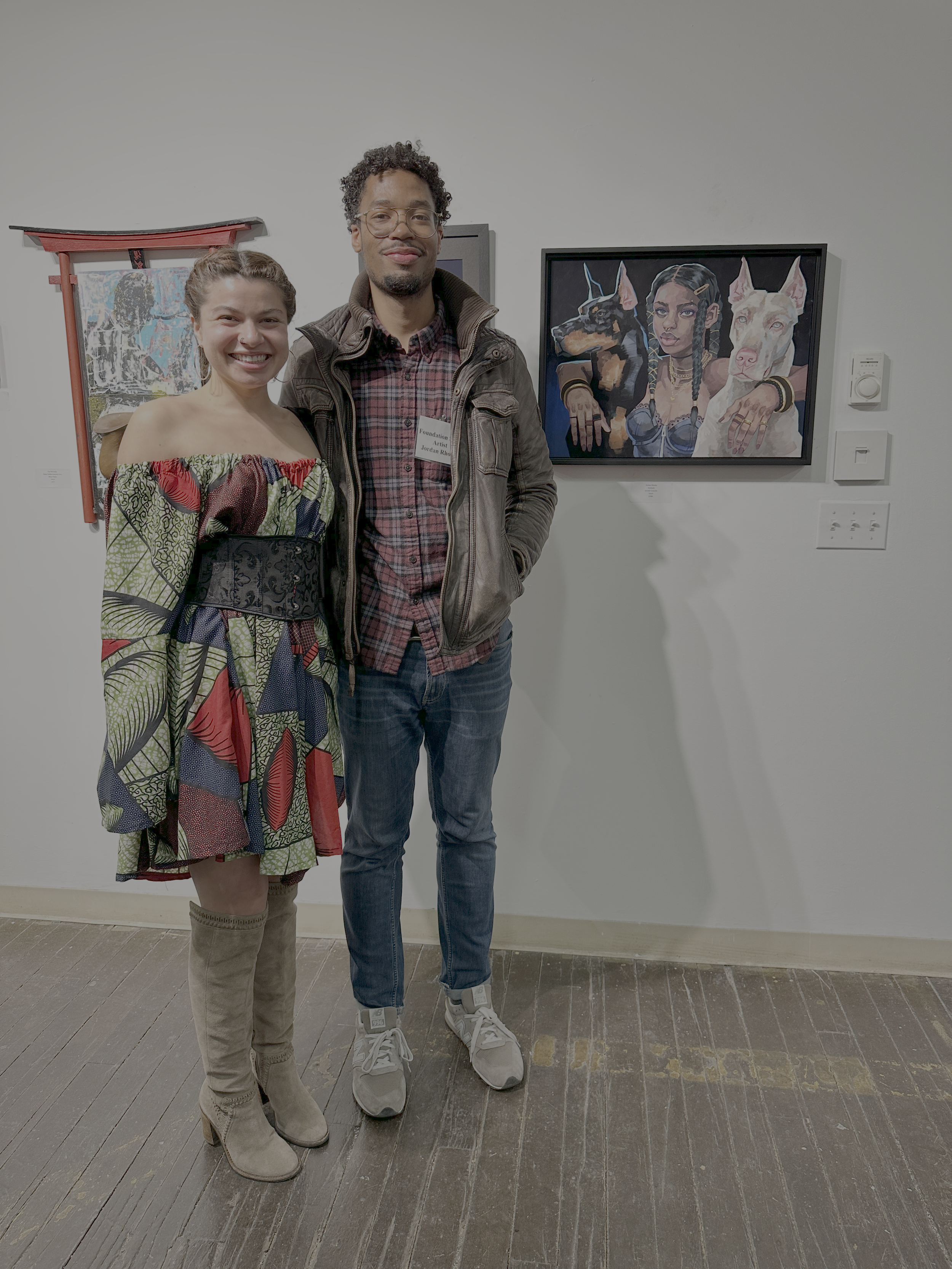 A woman and a man standing in an art gallery, smiling at the camera. The woman is wearing a colorful off-shoulder dress and the man is dressed casually in a plaid shirt, leather jacket, and jeans. Behind them is a digital painting featuring a woman with braided hair and two dogs.