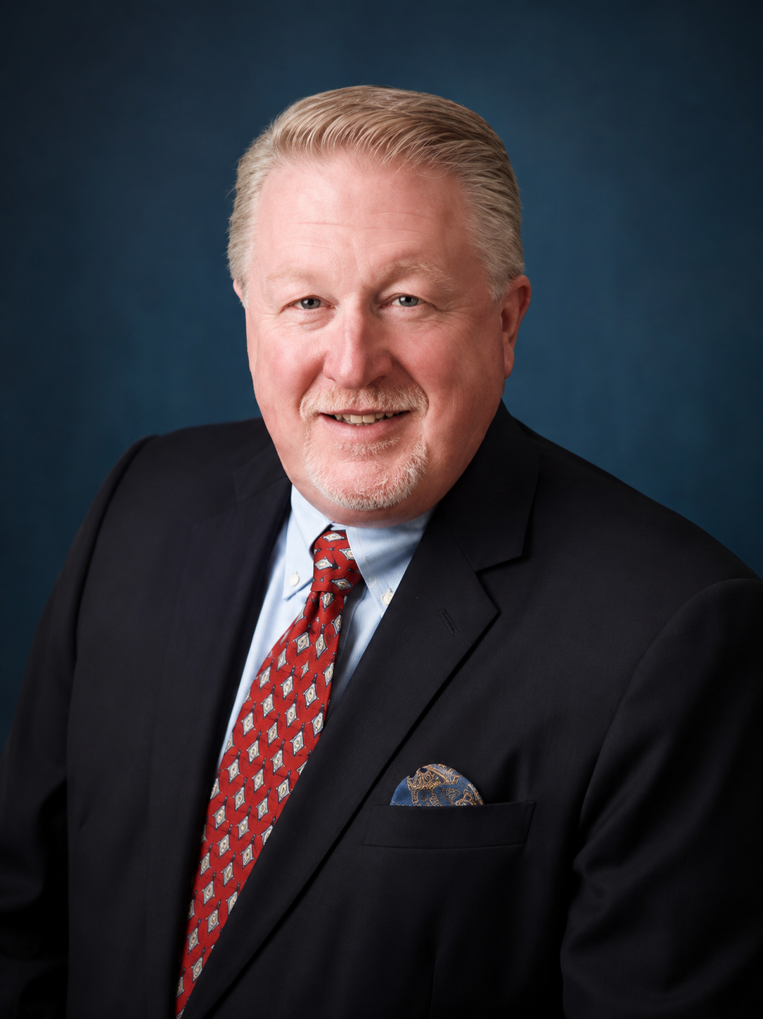 Portrait of a middle-aged man with light hair and a beard, wearing a dark suit, light blue shirt, and a red patterned tie, smiling against a dark blue background.