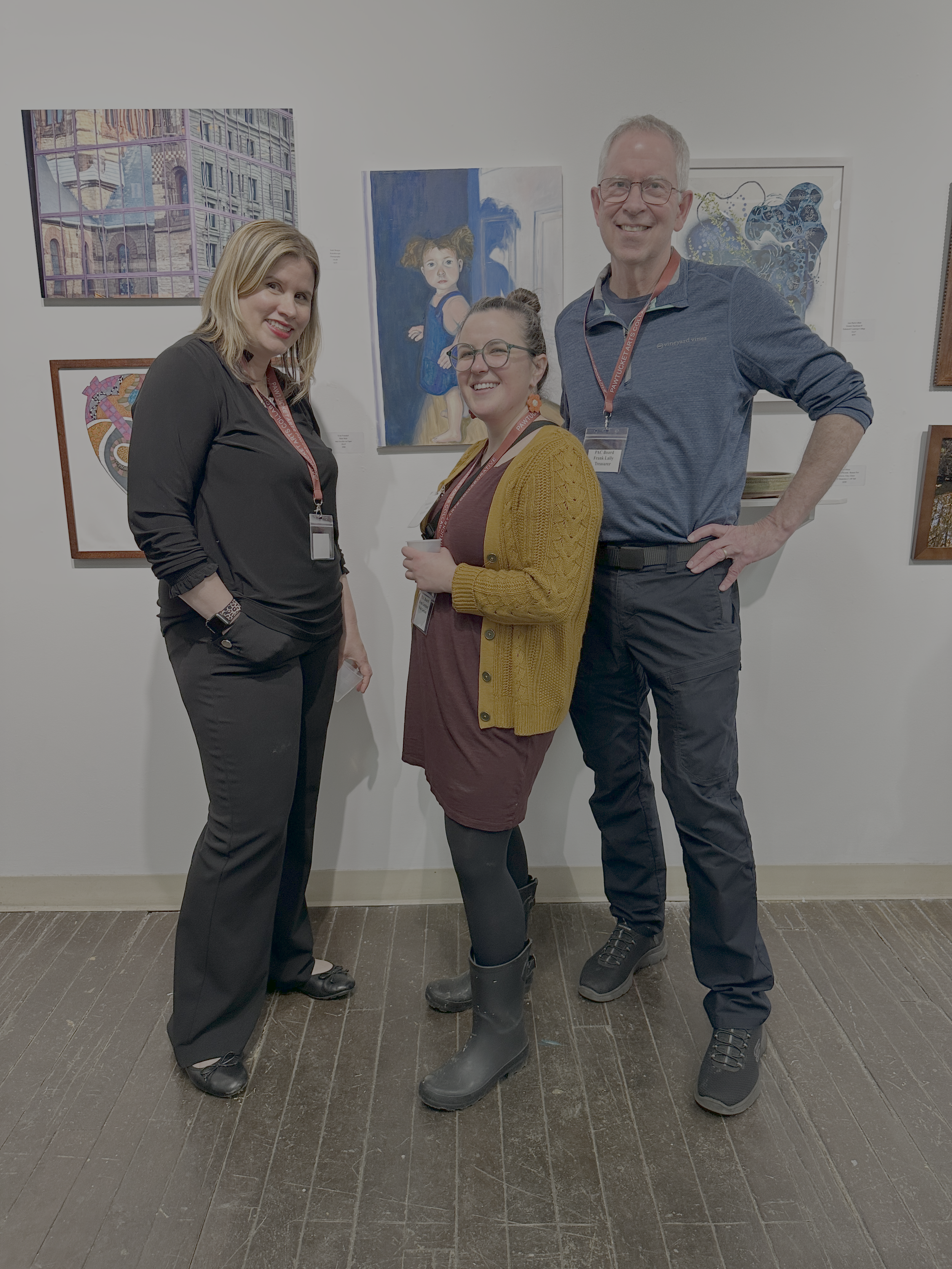 Three people standing in an art gallery, smiling at the camera. Two women and one man. The woman on the left has blonde hair and is wearing black pants and a black top. The woman in the middle has glasses, brown hair in a bun, and is wearing a mustard yellow cardigan over a brown dress. The man on the right has short gray hair, glasses, and is wearing a blue pullover, black pants, and black boots. Behind them are several colorful paintings on the white gallery wall.
