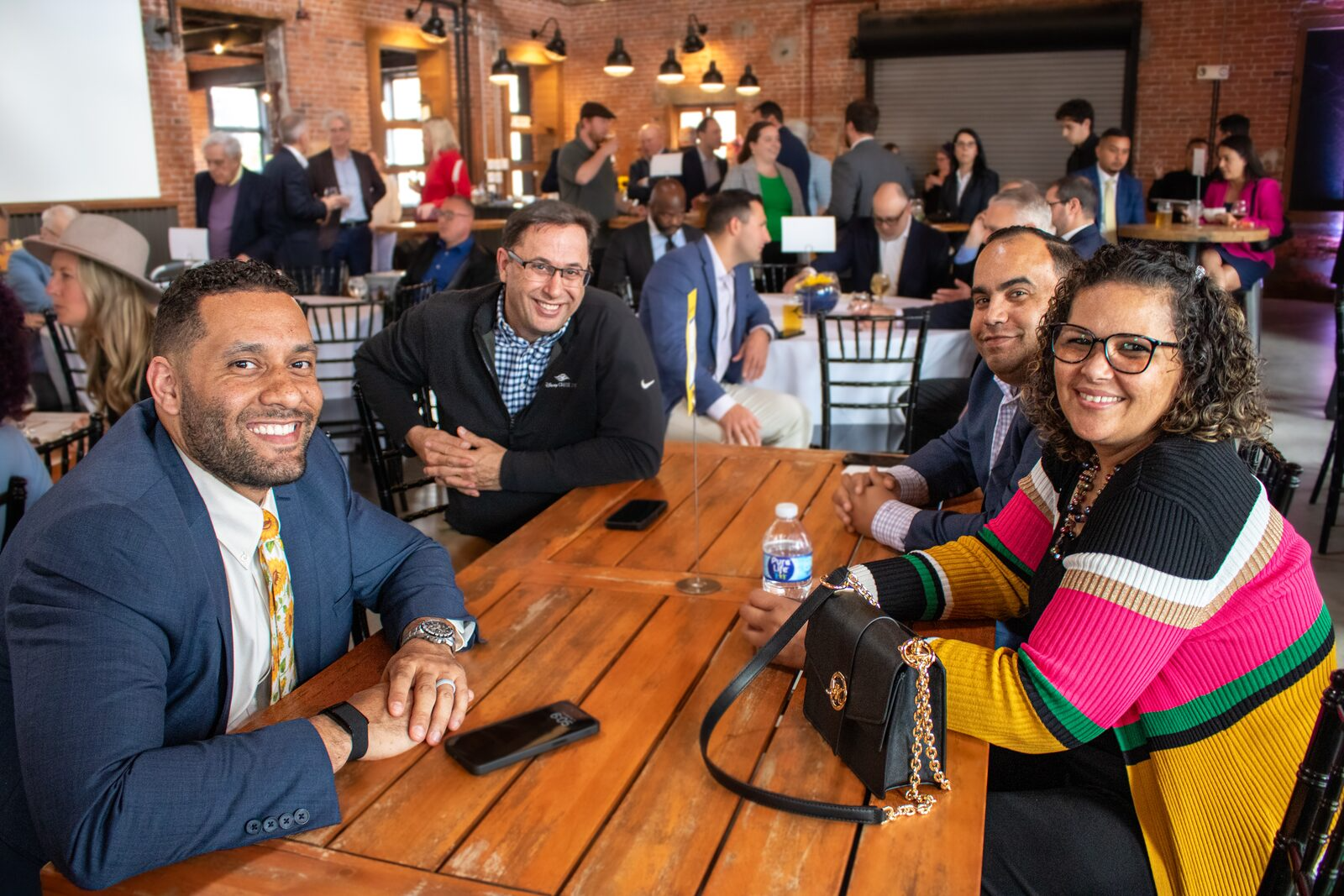 Four people seated at a wooden table in a busy, brick-walled event space, smiling at the camera. Behind them, other attendees are mingling and standing near tables, with warm lighting and windows in the background.