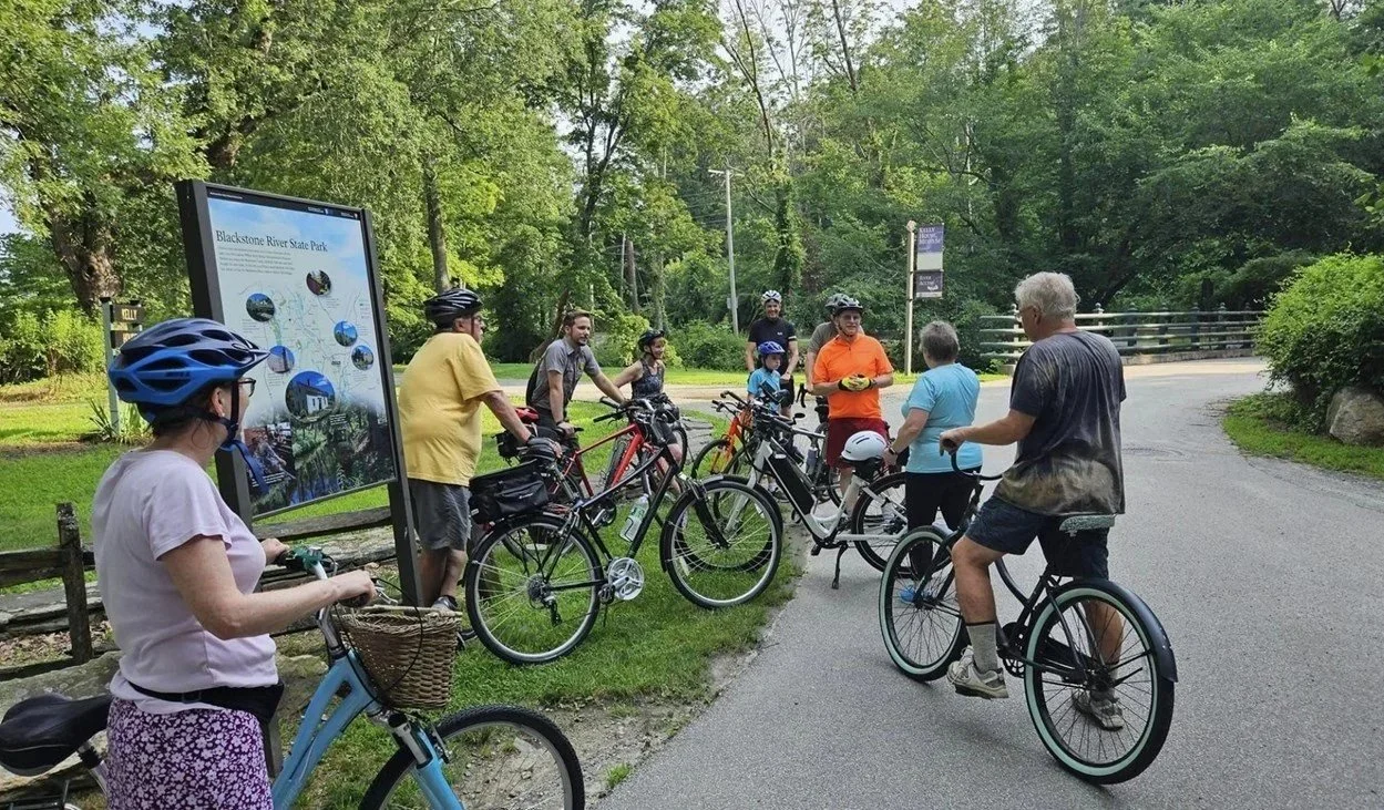 Group of people with bicycles gathered at a park entrance with a sign, surrounded by green trees.