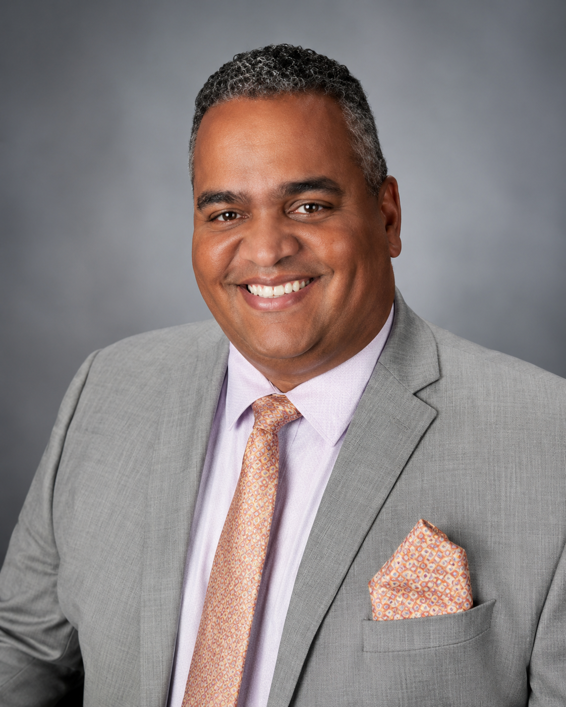 Professional headshot of a smiling man wearing a light gray suit, light purple dress shirt, light orange patterned tie, and matching pocket square against a gray background.