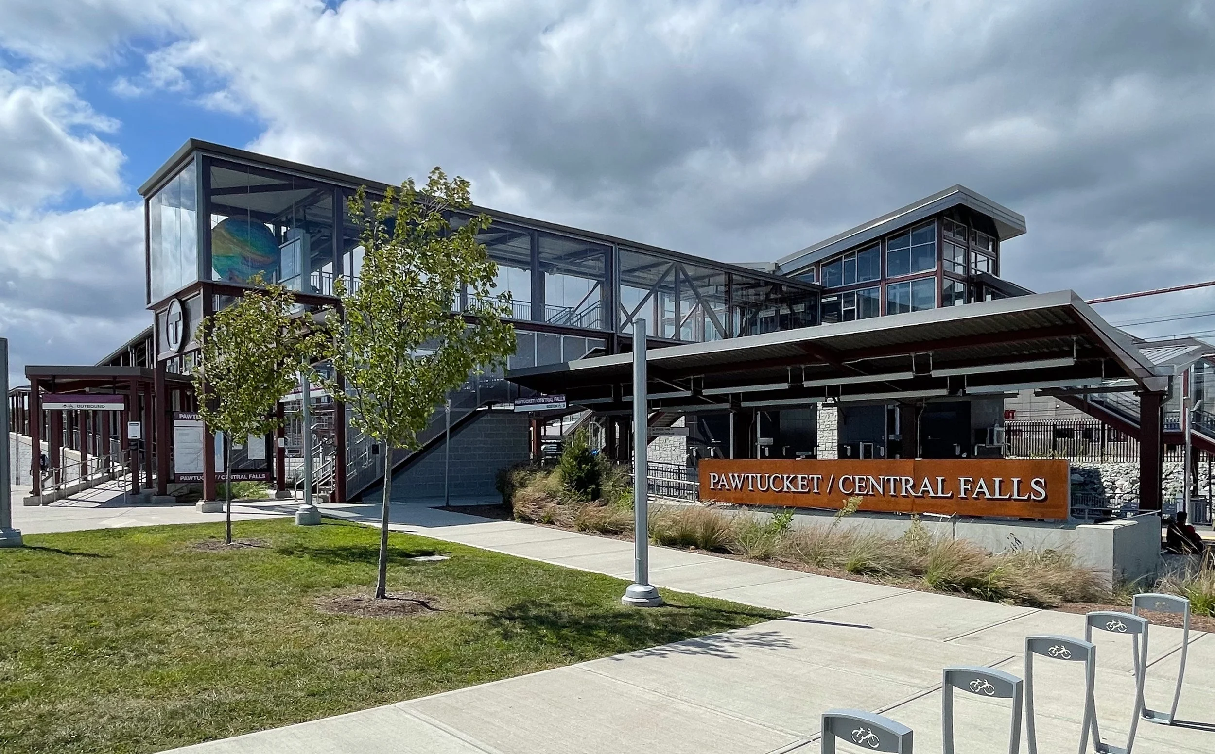 Modern transportation hub with glass-enclosed walkways, a sign reading 'Pawtucket / Central Falls,' green grass, walking paths, and a cloudy sky.