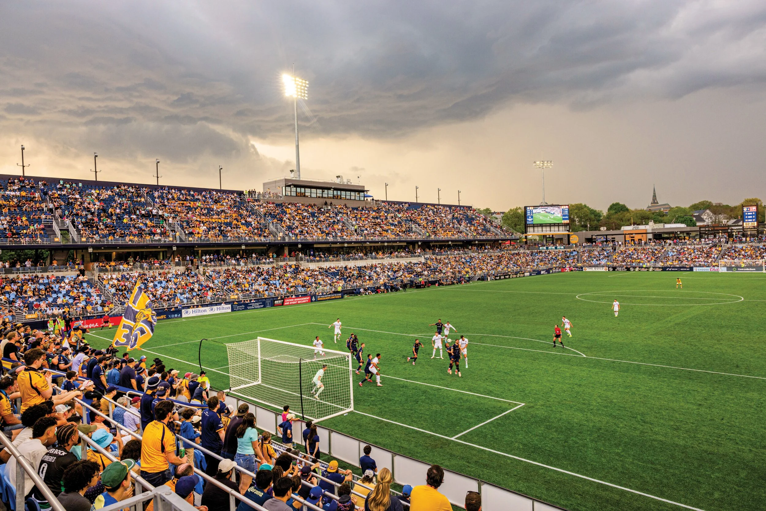 A soccer stadium filled with spectators, with players on the field and a cloudy sky overhead, illuminated by stadium lights.