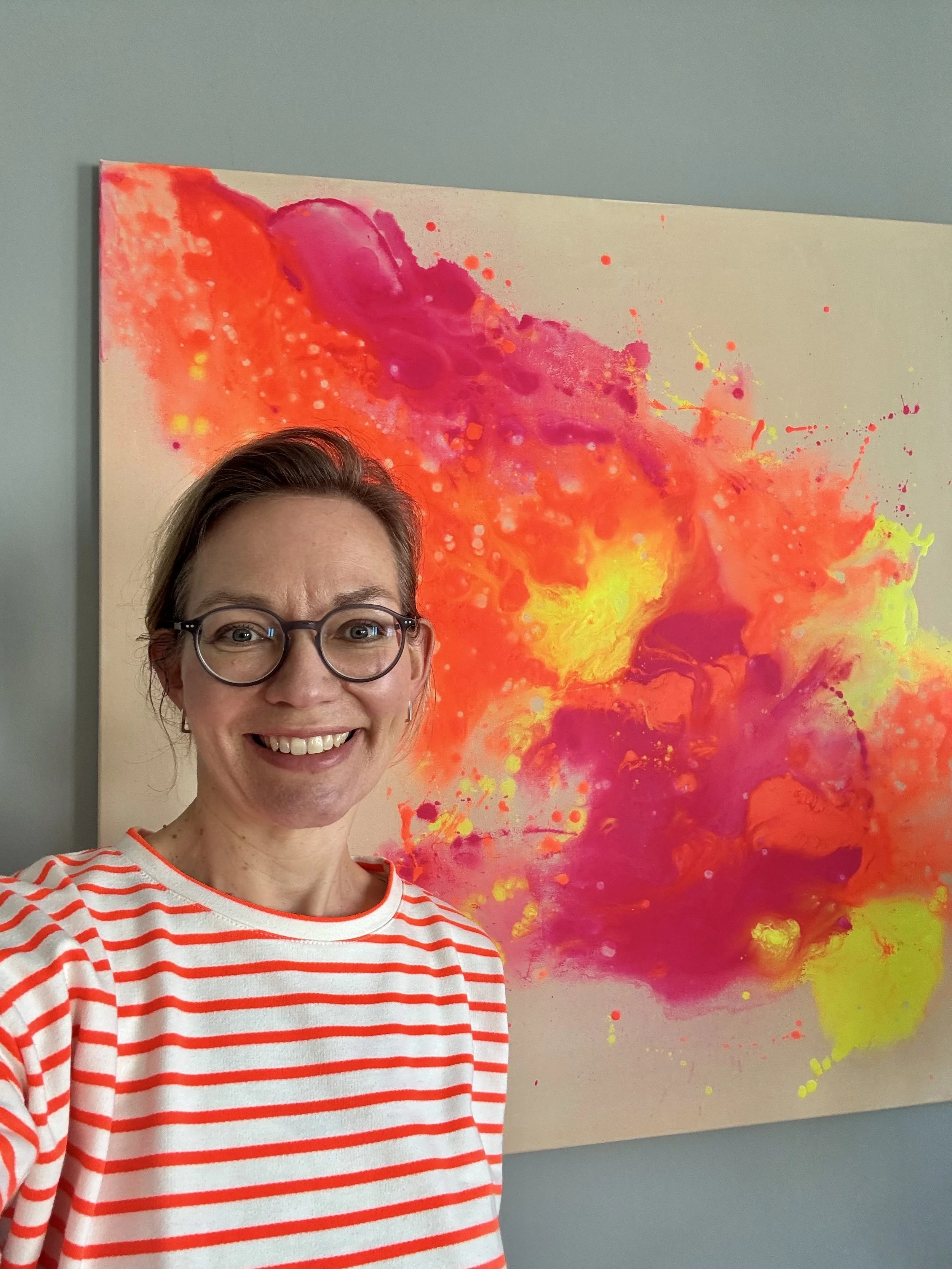 Eine lachende Frau mit Brille in einem weiß-roten gestreiften Hemd posiert vor einem bunten abstrakten Gemälde mit Rosa, Gelb, Orange und Lila Farben.